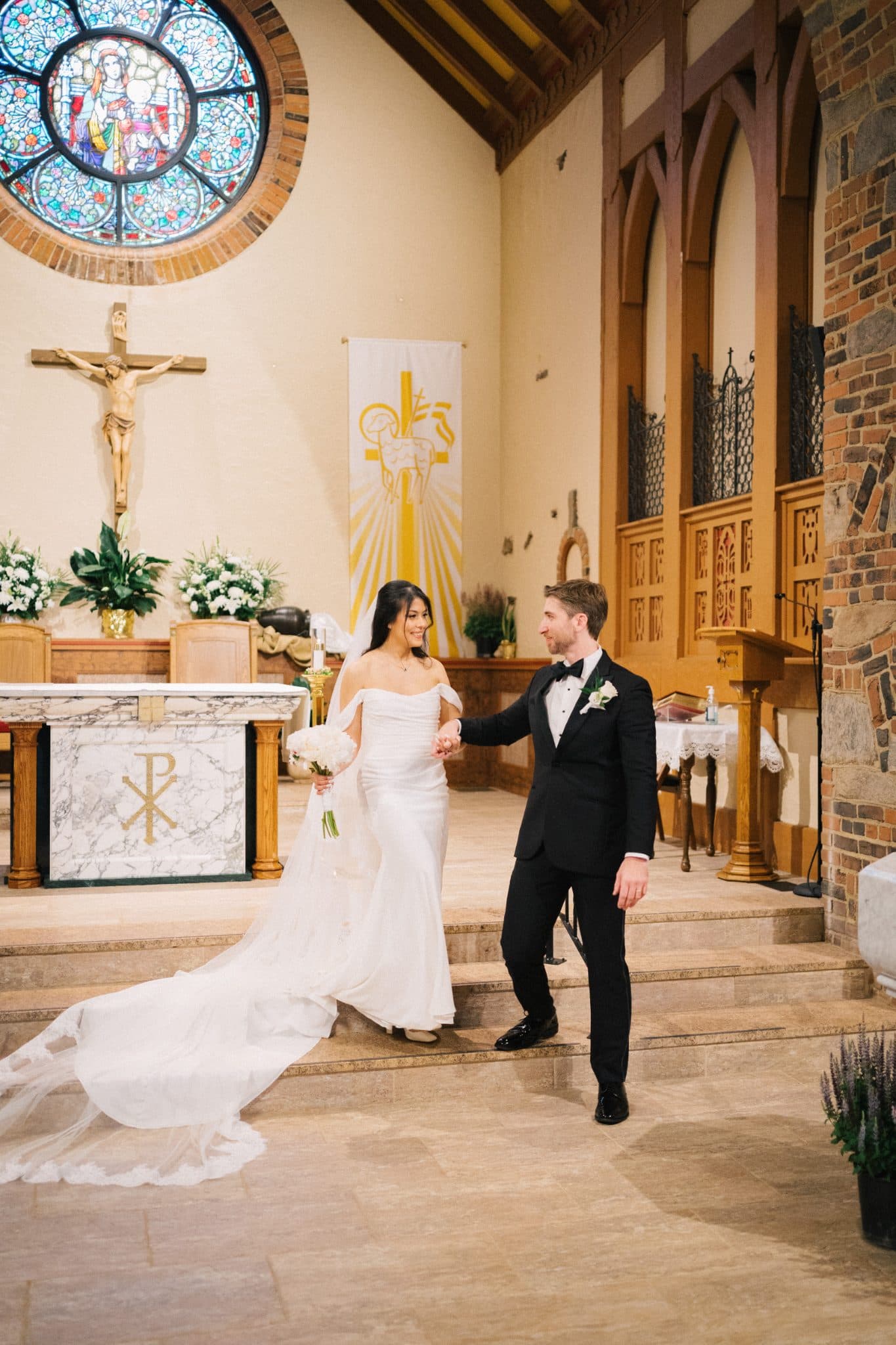 A bride in a white gown and a groom in a black tuxedo hold hands as they walk down the aisle in a church.