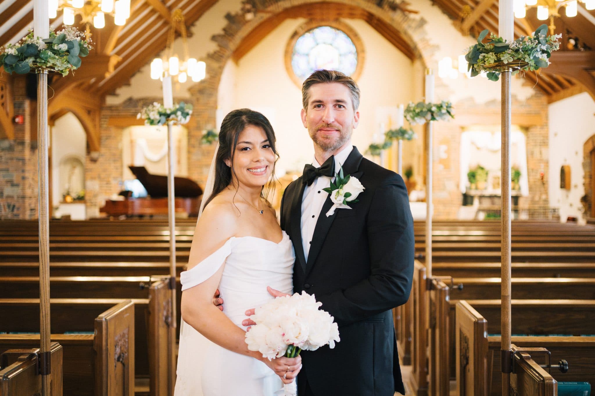 A bride in a white dress and a groom in a black suit stand together in a church, holding hands and smiling.