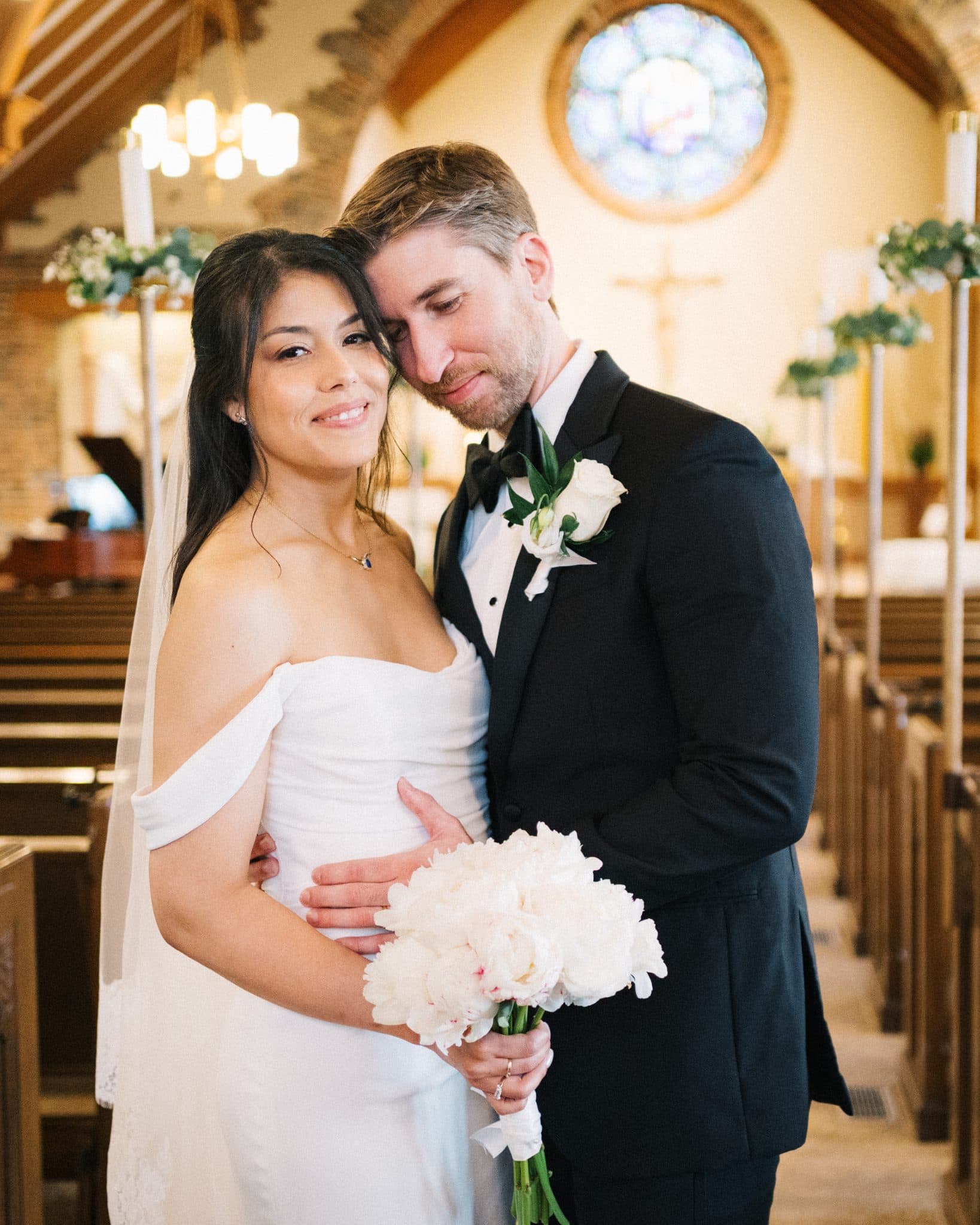 A bride in a white dress and veil holds a bouquet of white flowers, embracing a groom in a black suit with a boutonniere, in a church setting with pews and stained…