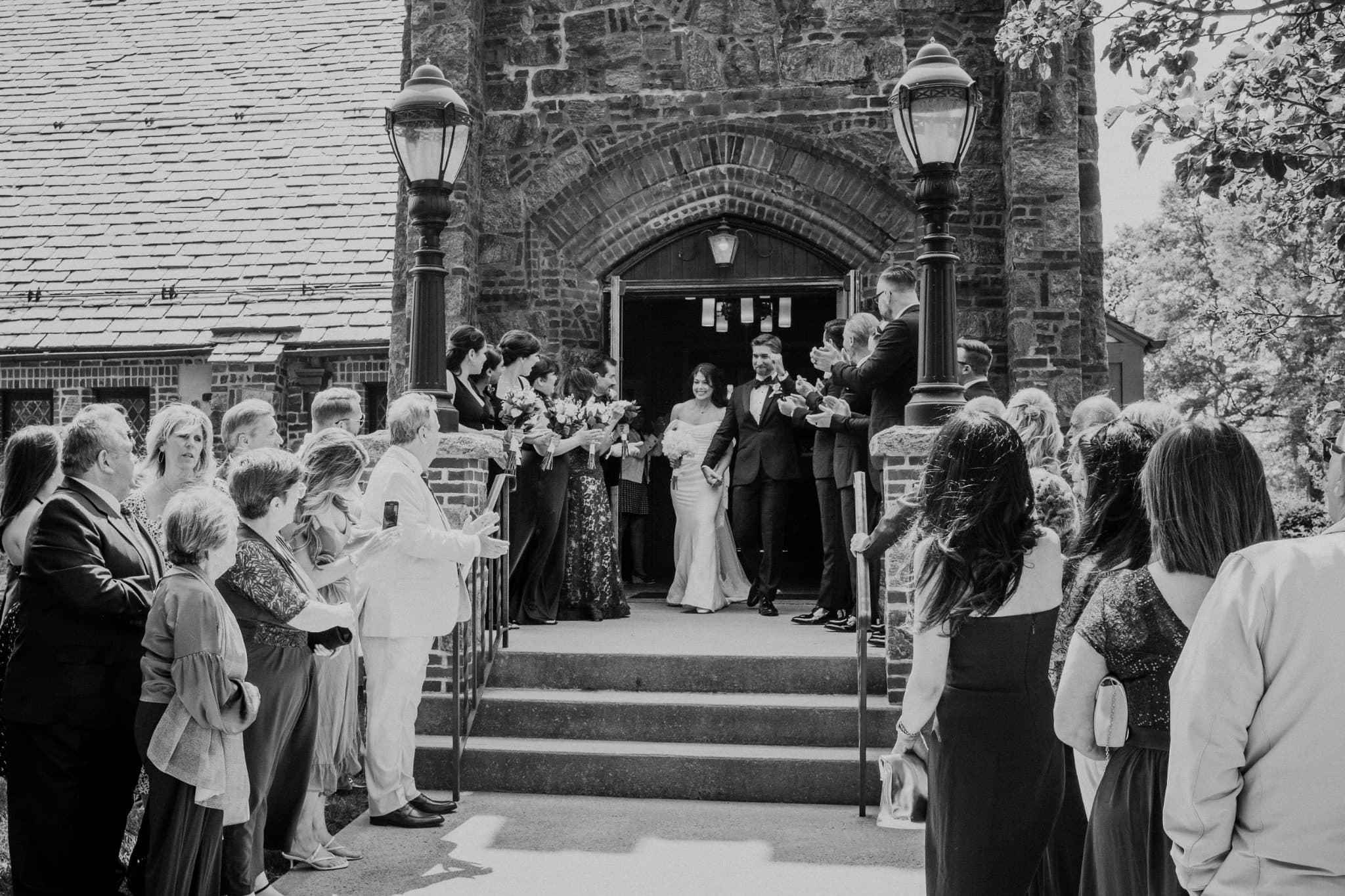A bride and groom exit a stone church, greeted by guests clapping and smiling.