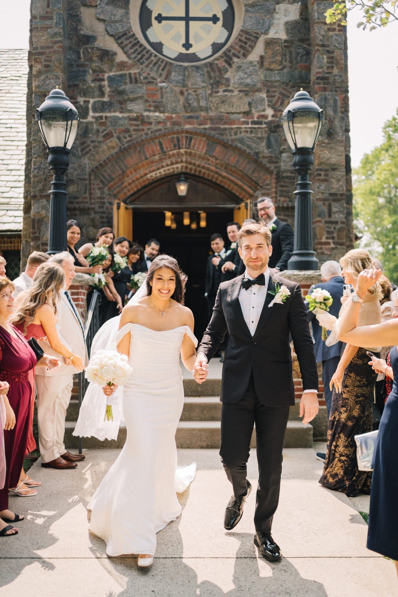 A bride in a white off-shoulder gown and a groom in a black tuxedo walk hand in hand down a church aisle.