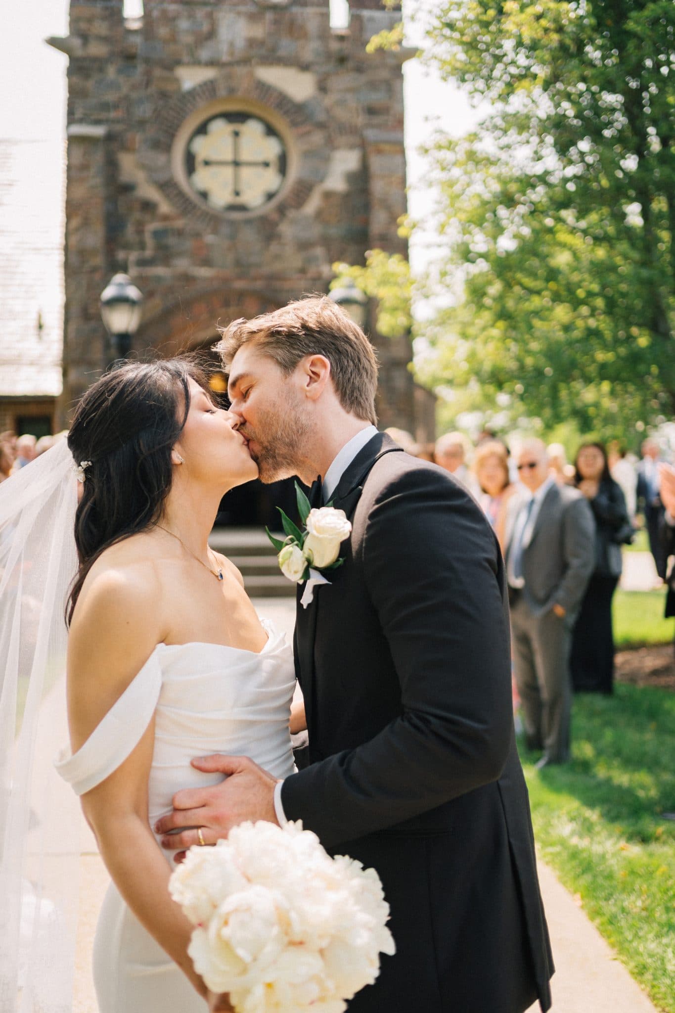 A bride in a white dress and veil kisses a groom in a black suit with a white boutonniere, holding a bouquet of white flowers, in front of a stone church with…