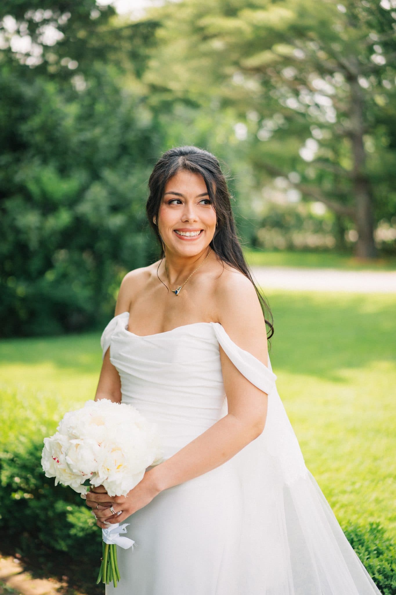 A bride in a white off-the-shoulder wedding dress holds a bouquet of white flowers, smiling and looking to the side.