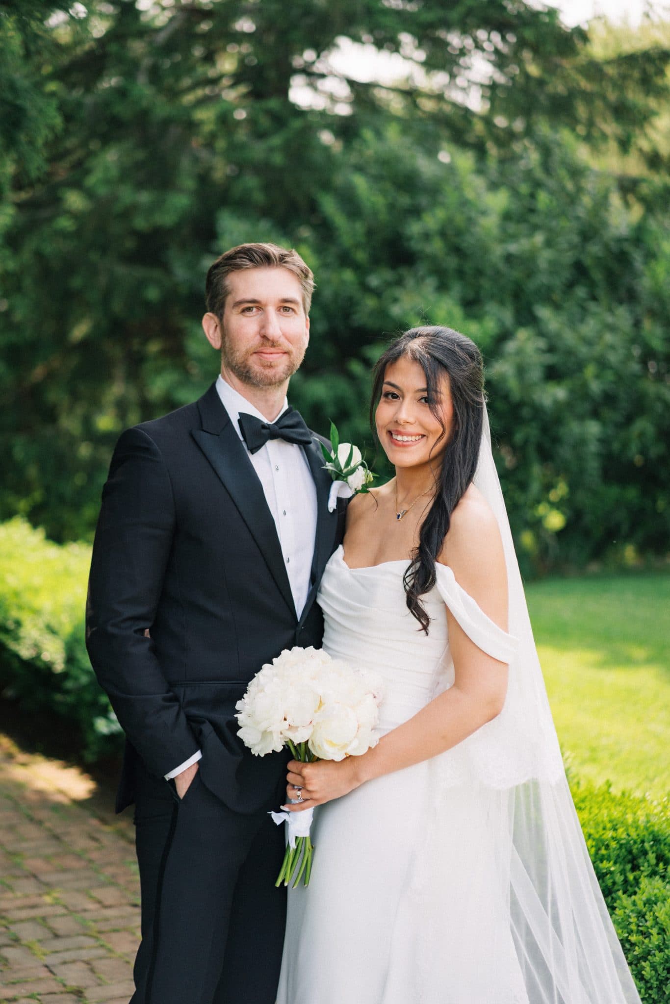 A bride in a white dress holds a bouquet of white flowers, standing beside a man in a black tuxedo with a bow tie.