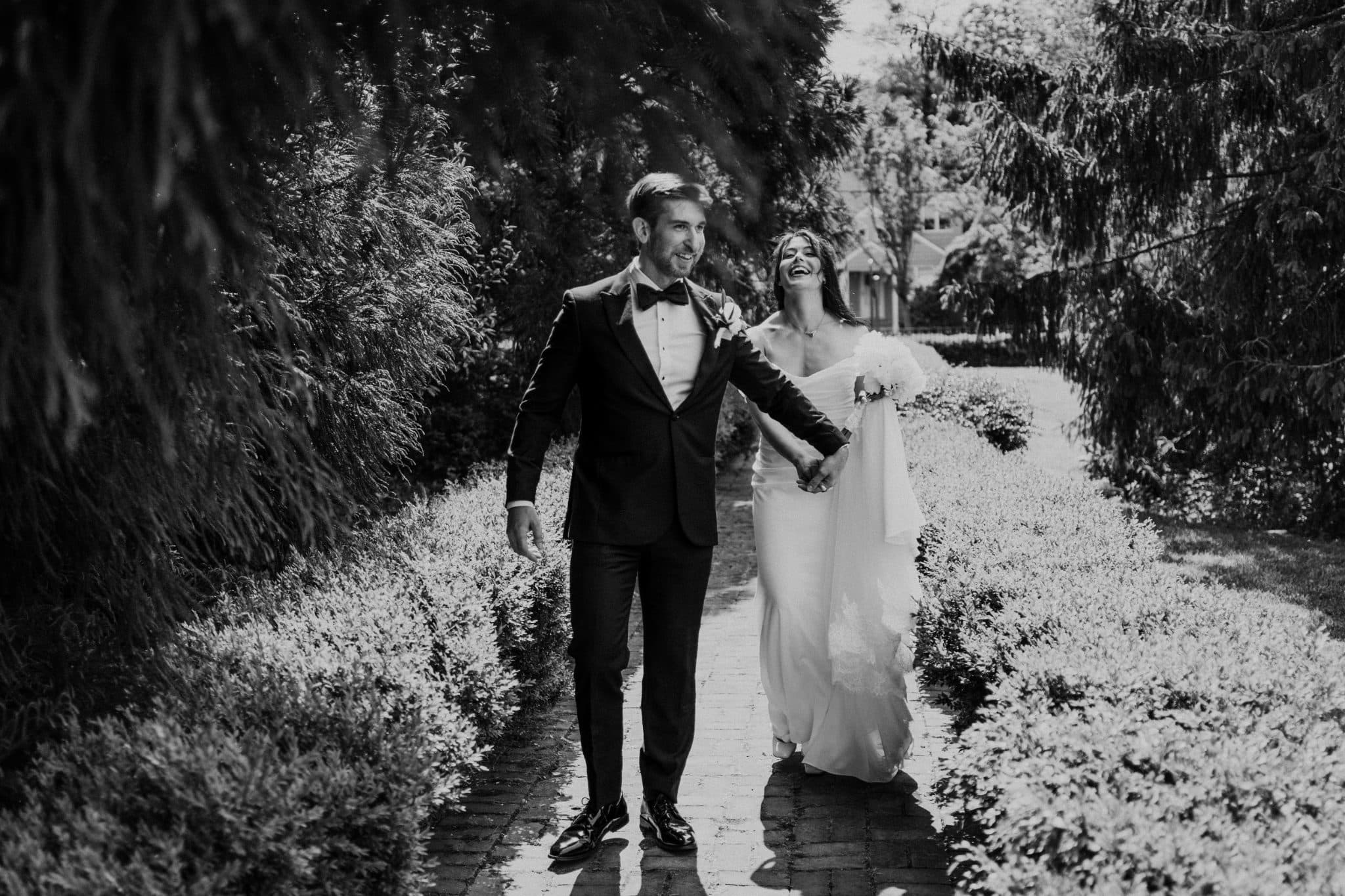 A bride in a white gown and a groom in a tuxedo walk hand in hand down a brick path lined with bushes.