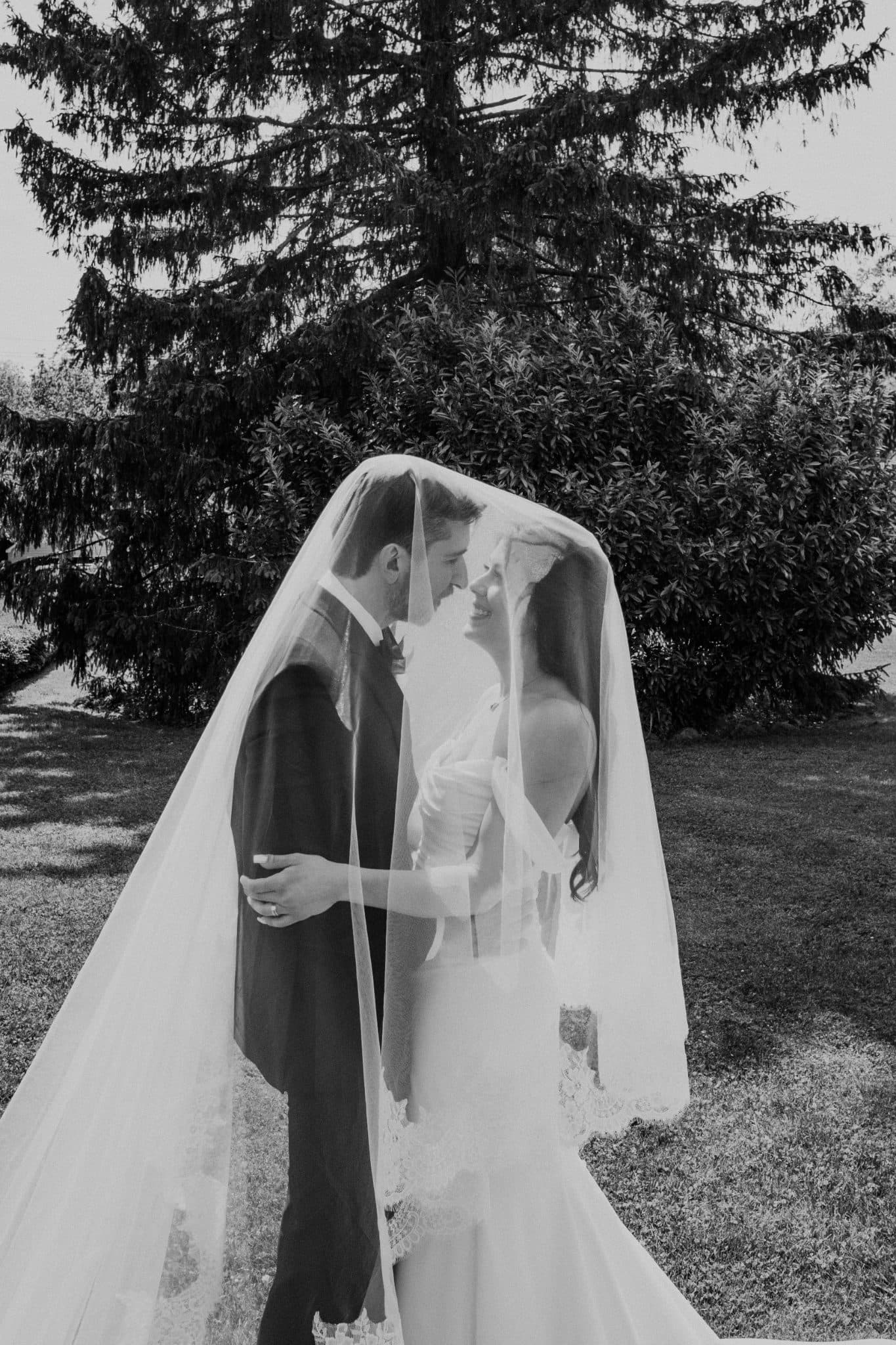 A bride and groom stand under a veil, embracing in a grassy area with trees in the background.