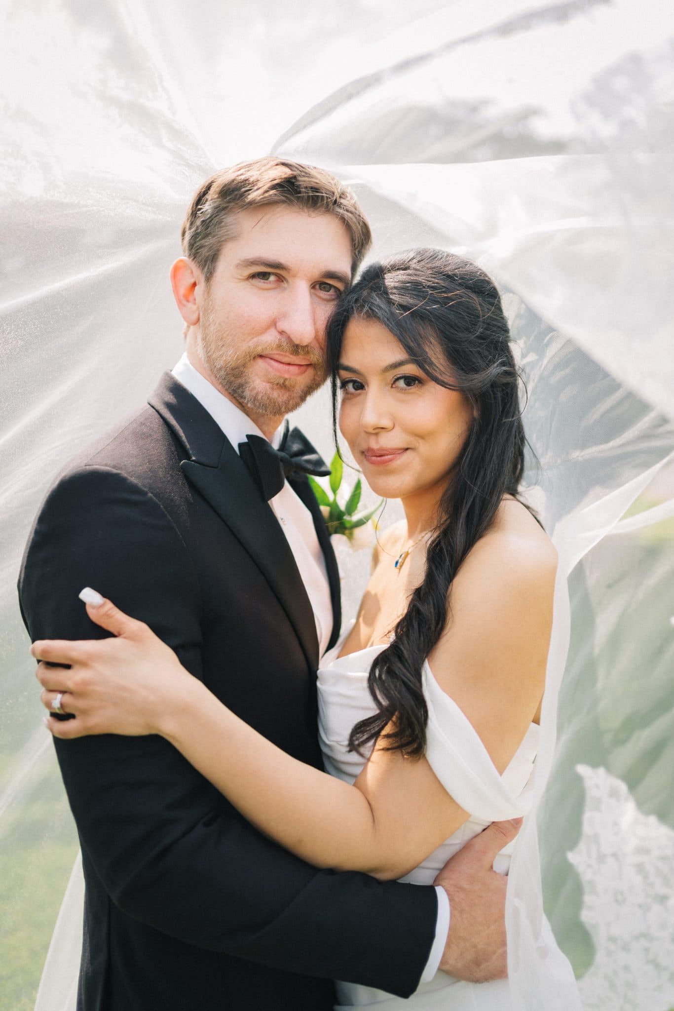 A couple embraces under a flowing white veil, the bride in a white dress and the groom in a black suit.