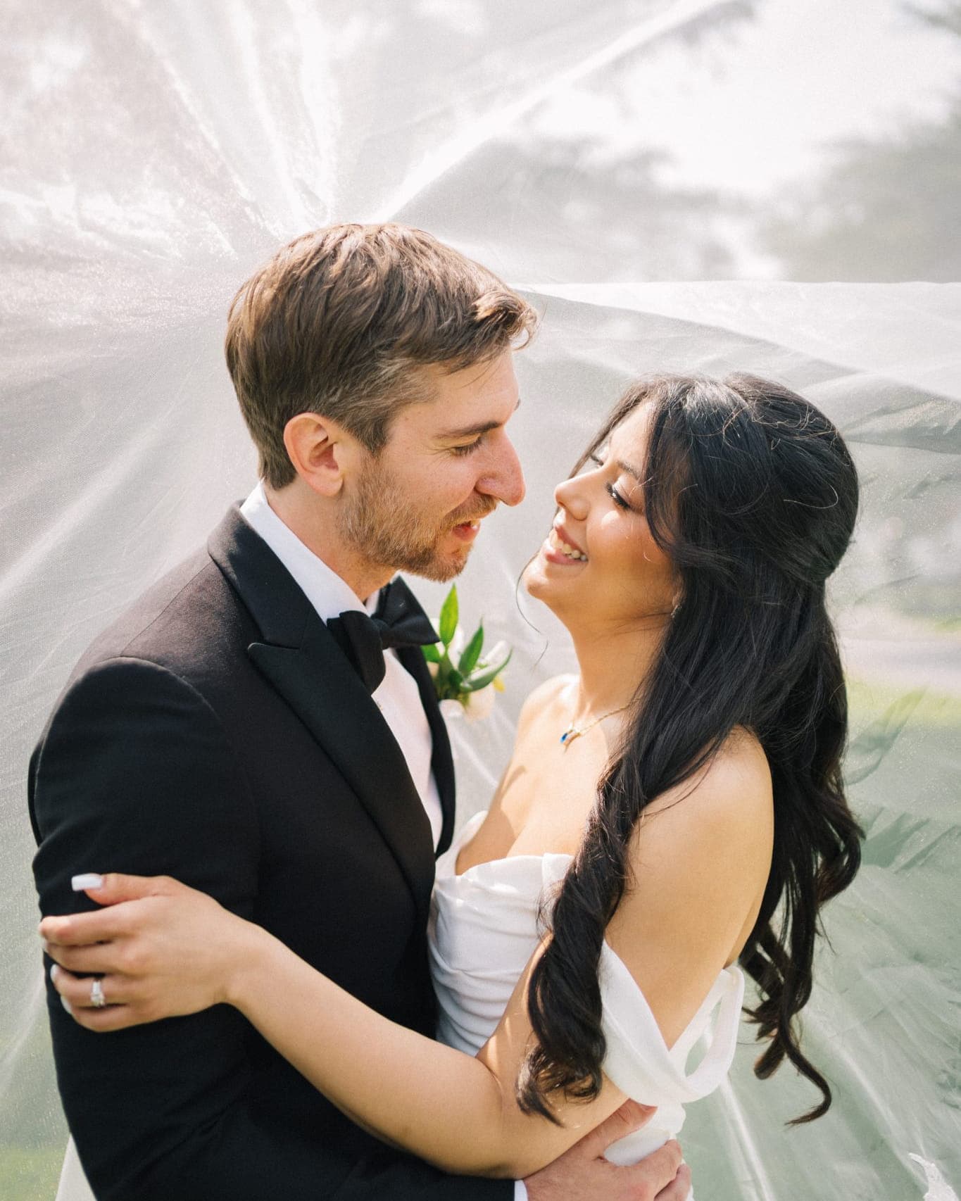 A bride in a white dress and a groom in a black suit embrace under a white veil.