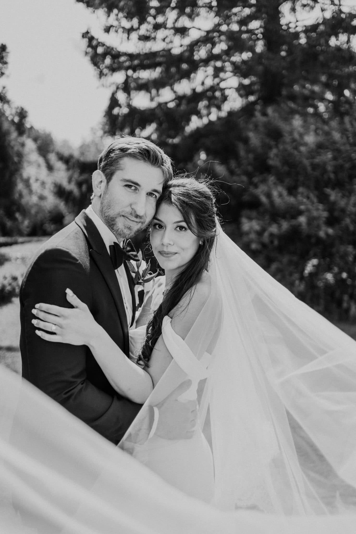 A bride and groom embrace under a flowing veil, surrounded by trees.