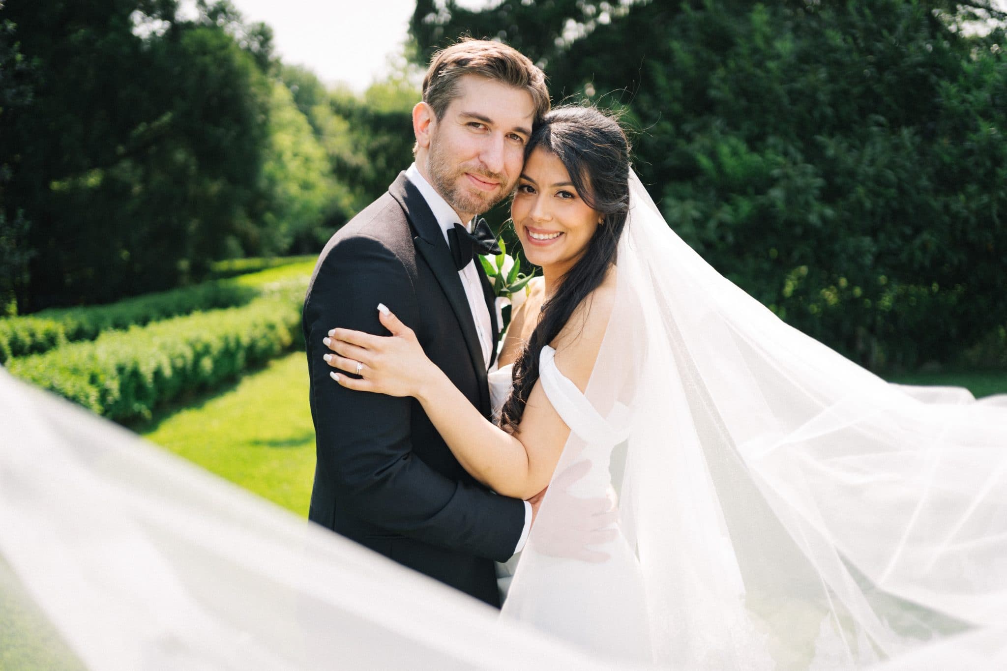A bride in a white dress and veil hugs a groom in a black suit against a backdrop of greenery.