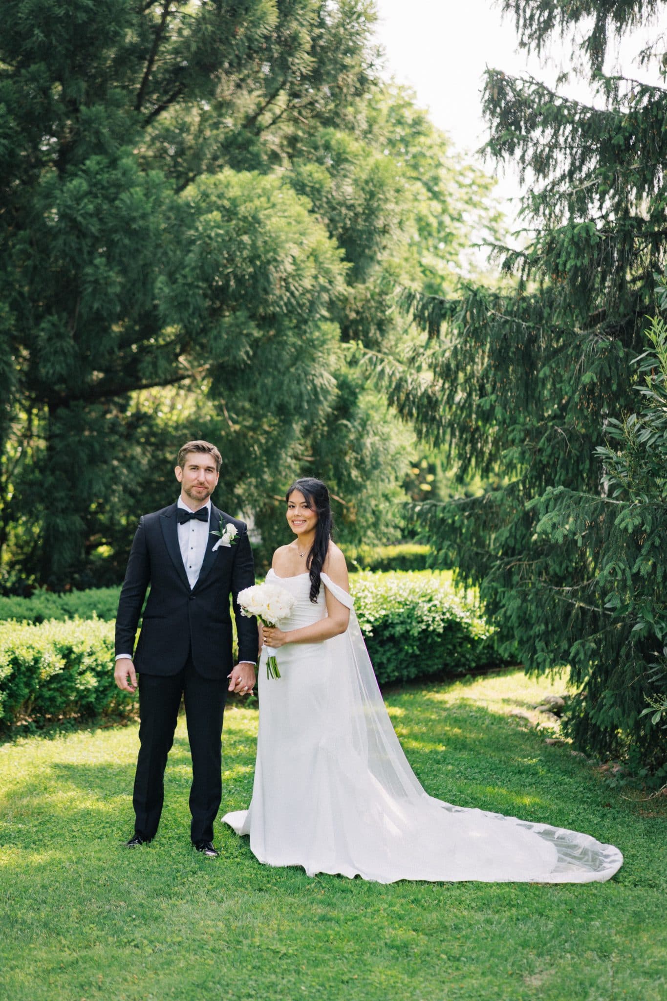 A bride in a white gown and a groom in a black tuxedo stand on a grassy lawn, holding hands, with trees in the background.