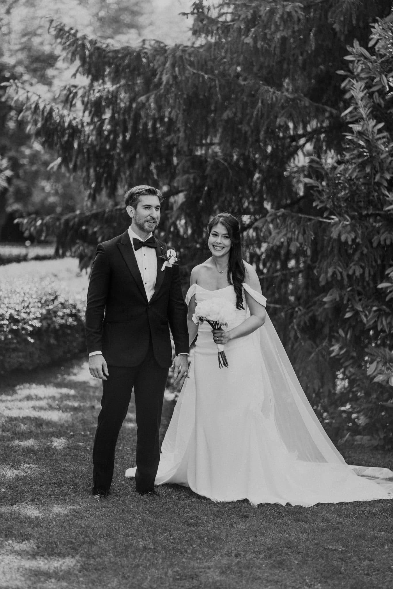 A bride in a white gown holds a bouquet, walking alongside a man in a black suit with a bow tie, surrounded by greenery.
