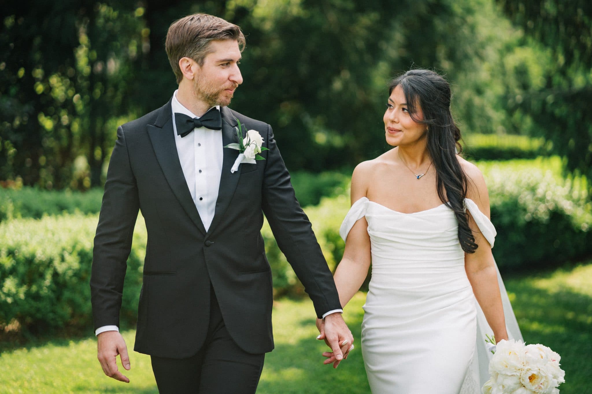 A couple in wedding attire holds hands, walking in a lush green garden.
