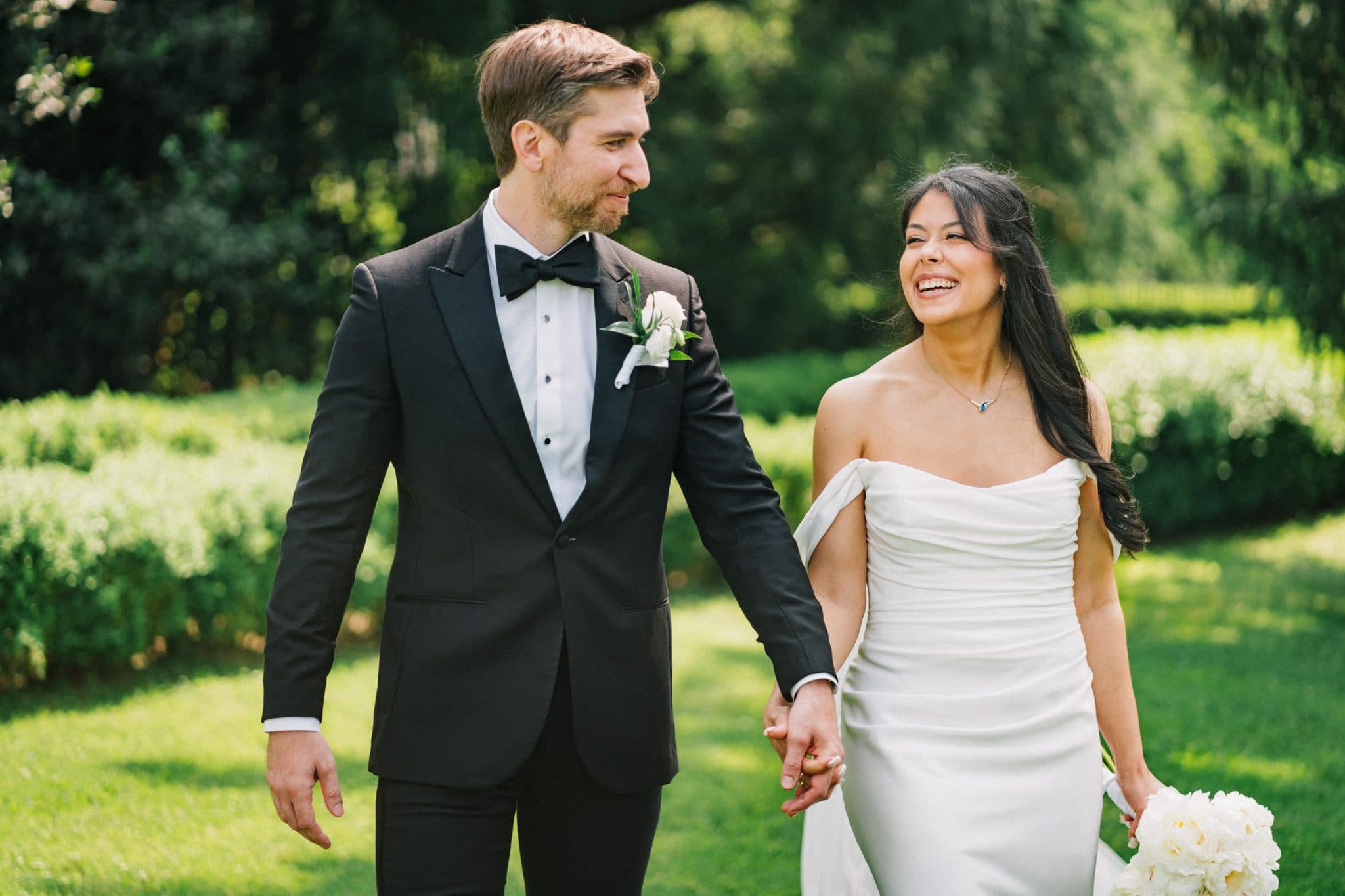 A couple in wedding attire holds hands, walking through a lush green garden.