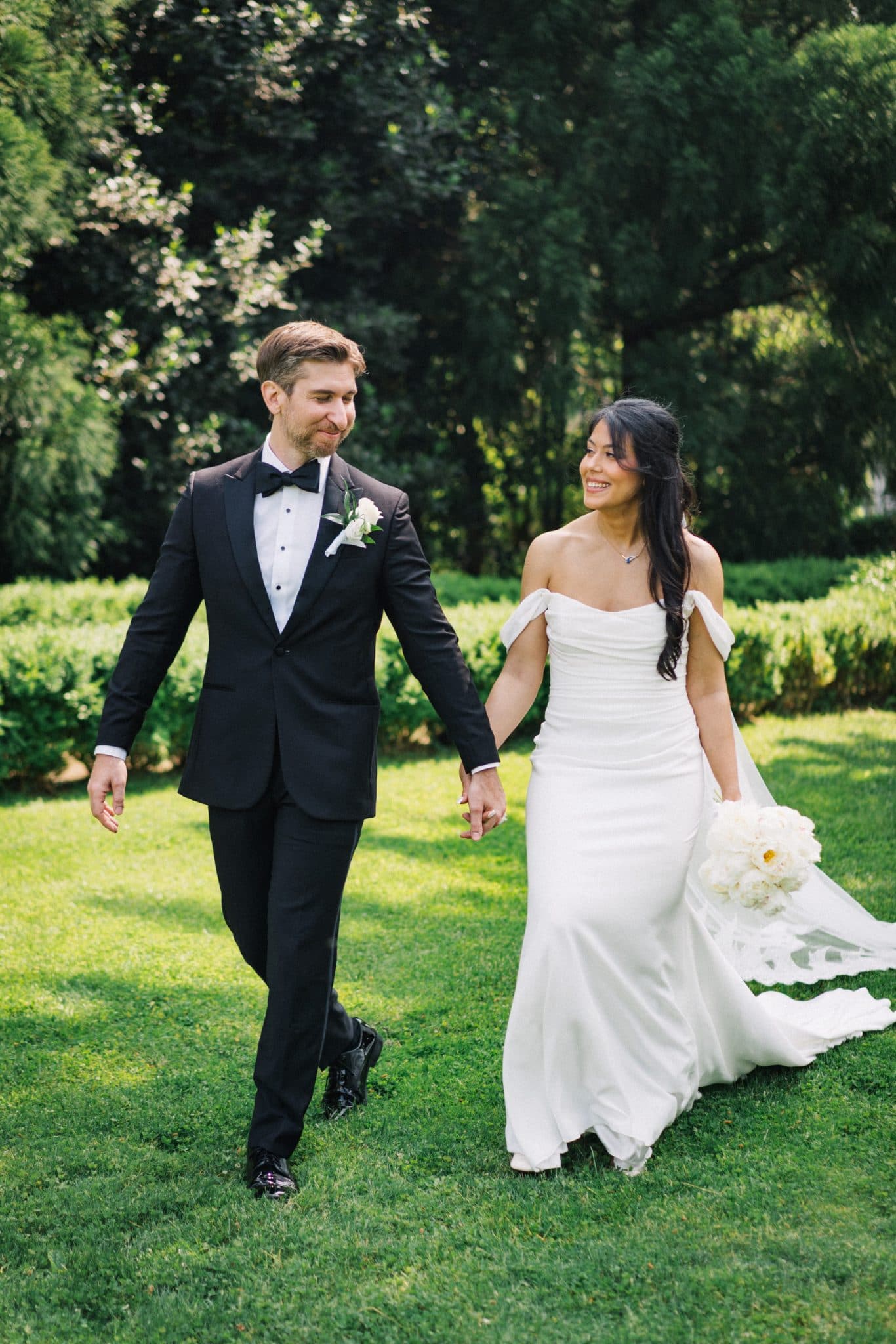 A couple in wedding attire walks hand in hand on a grassy lawn, surrounded by trees.
