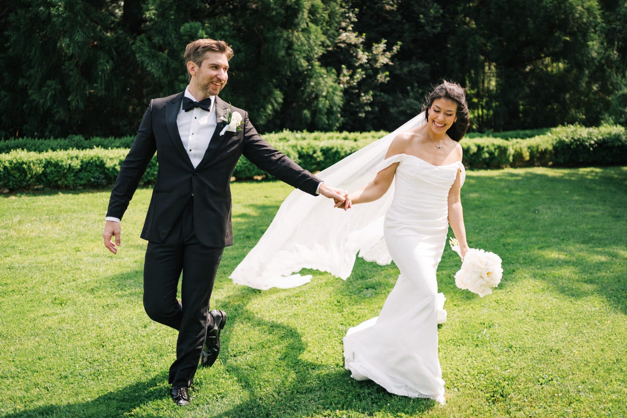 A bride in a white off-shoulder gown and a groom in a black tuxedo hold hands, walking on a grassy lawn with a bouquet of white flowers.