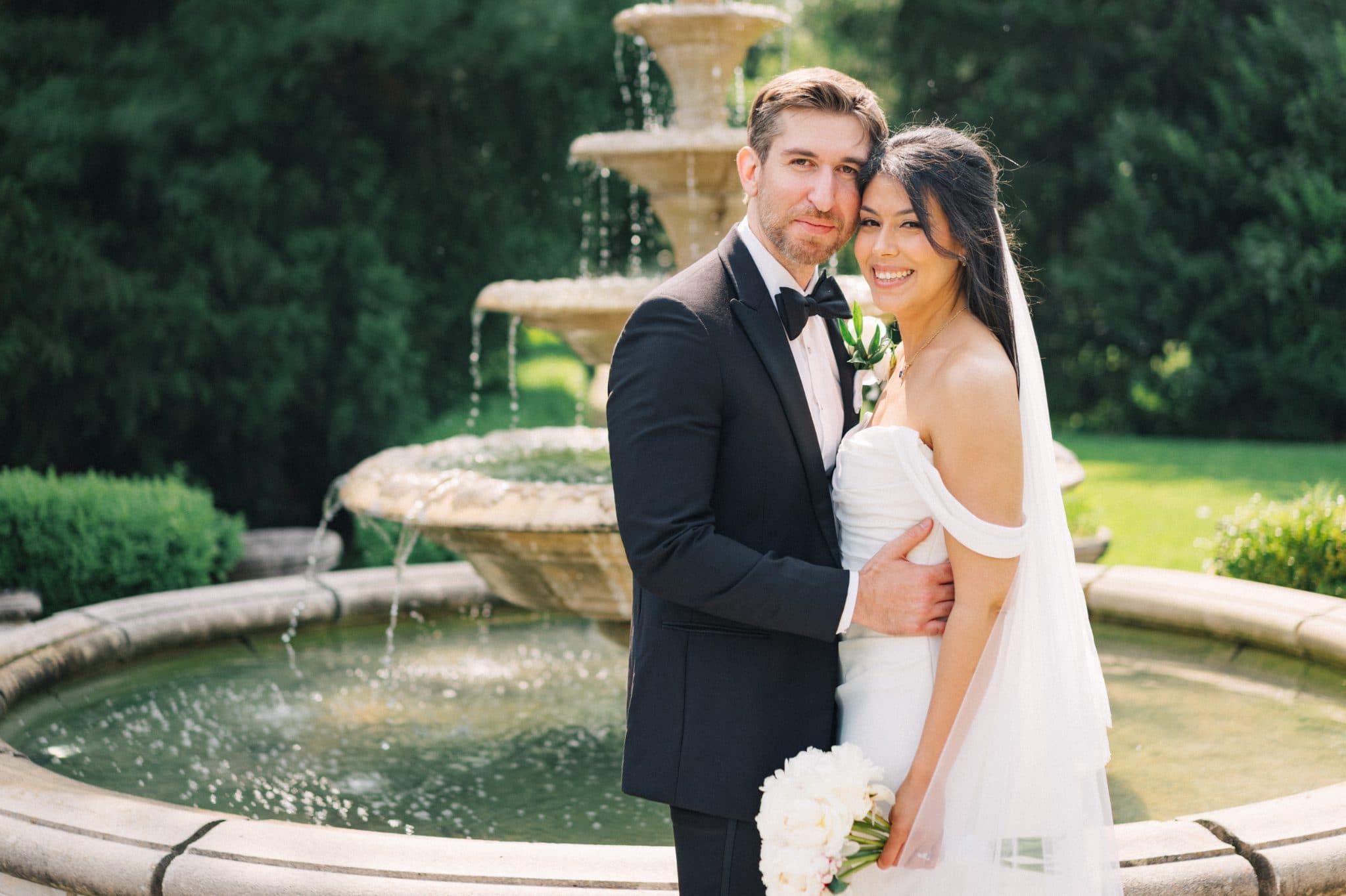 A bride in a white dress and veil stands beside a groom in a black suit, both smiling, with a fountain in the background.