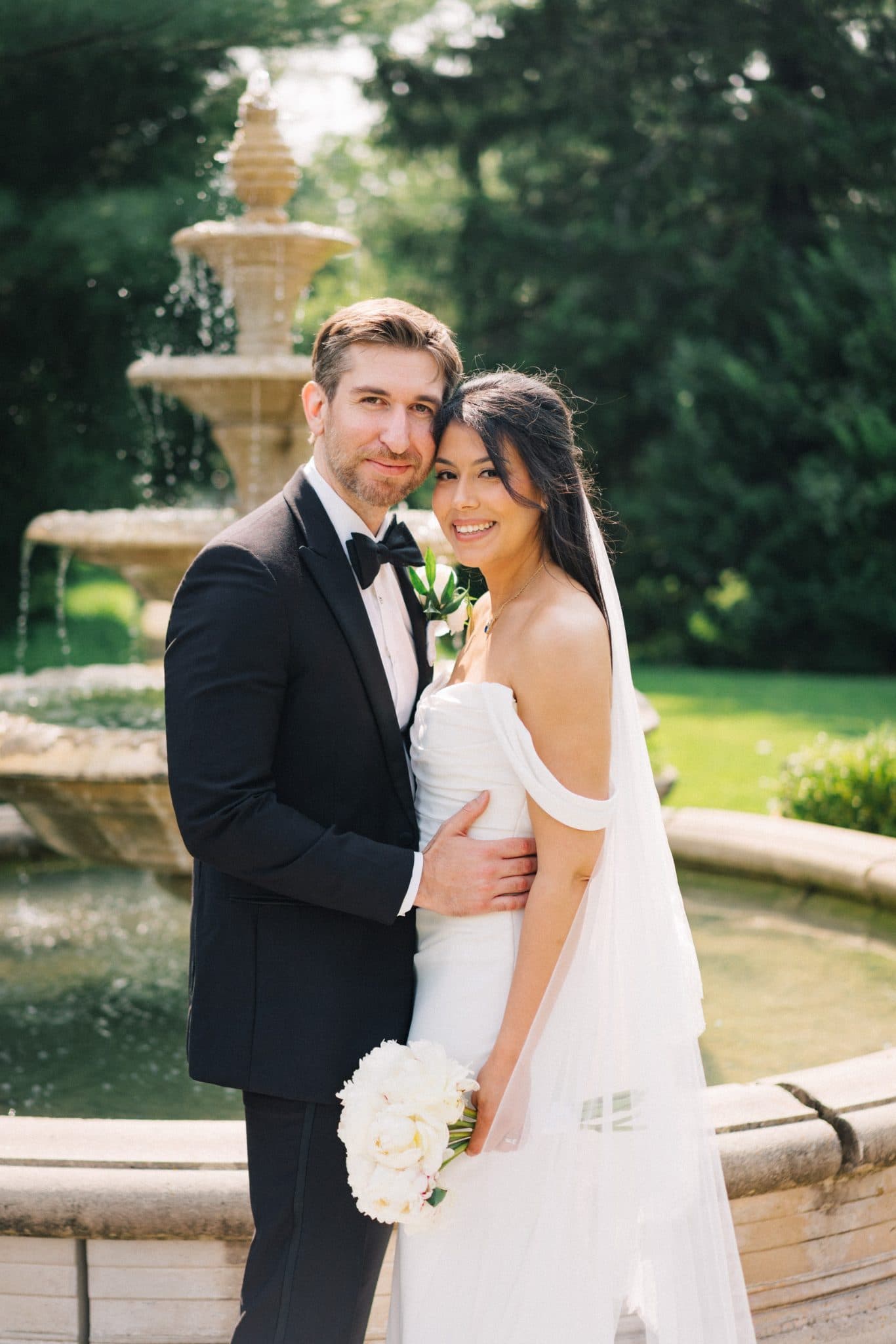 A bride in a white dress holds a bouquet of white flowers, standing beside a groom in a black tuxedo, with a fountain and lush greenery in the background.