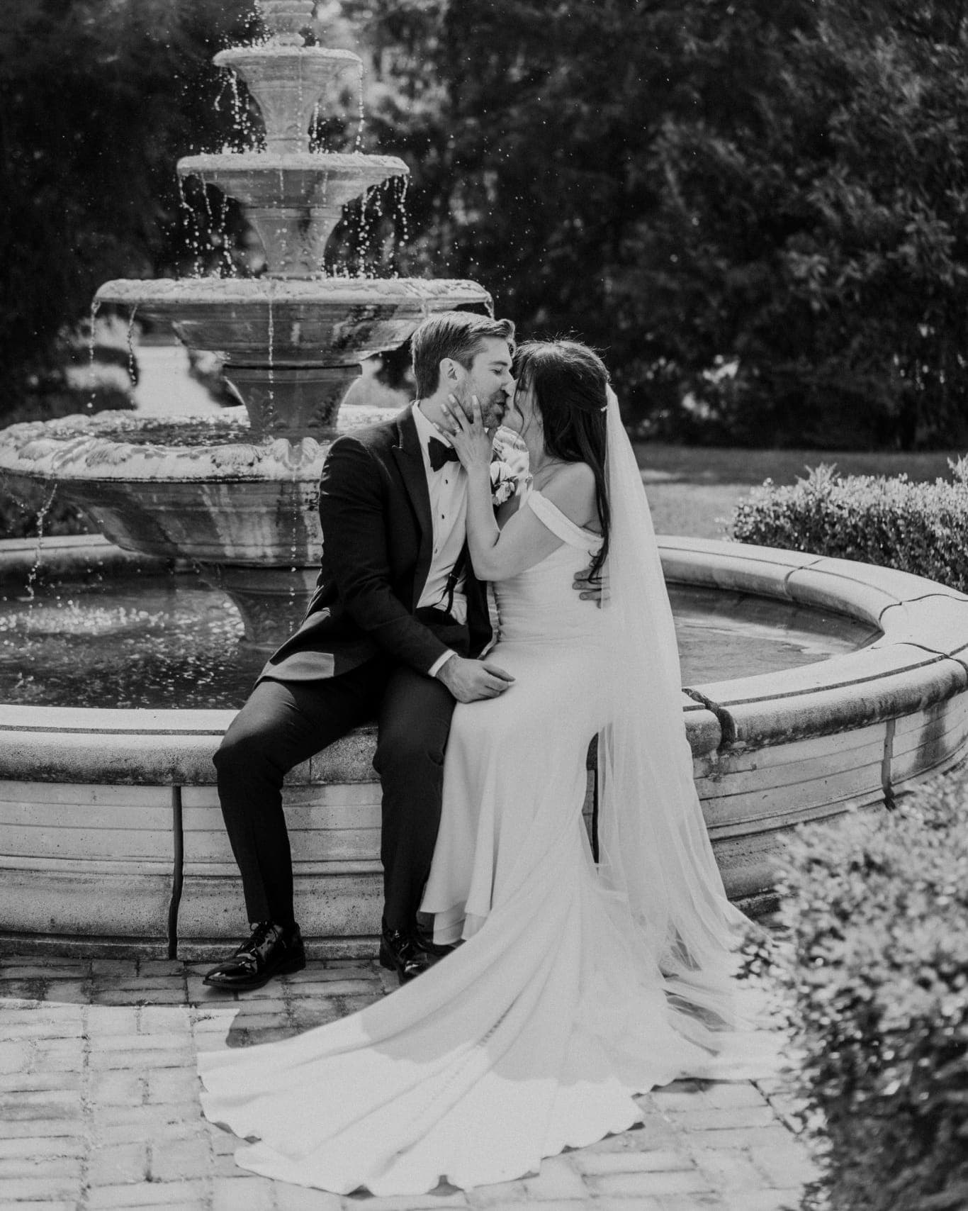 A bride and groom sit on a fountain ledge, the bride in a flowing white gown and veil, the groom in a dark suit, both facing each other.
