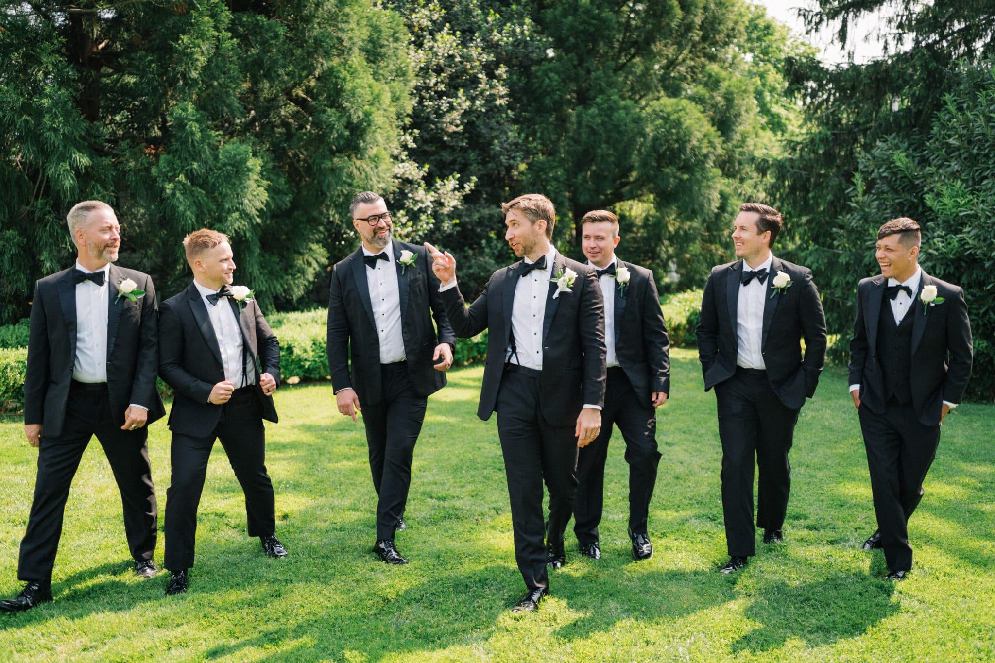 A group of men in black tuxedos and bow ties stand on a grassy lawn, smiling and interacting with each other.