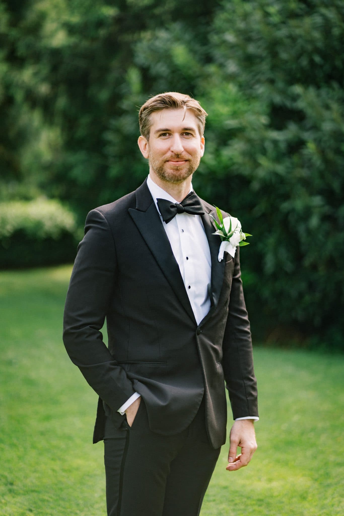 A man in a black tuxedo with a white shirt and black bow tie stands outdoors with a boutonniere on his lapel.