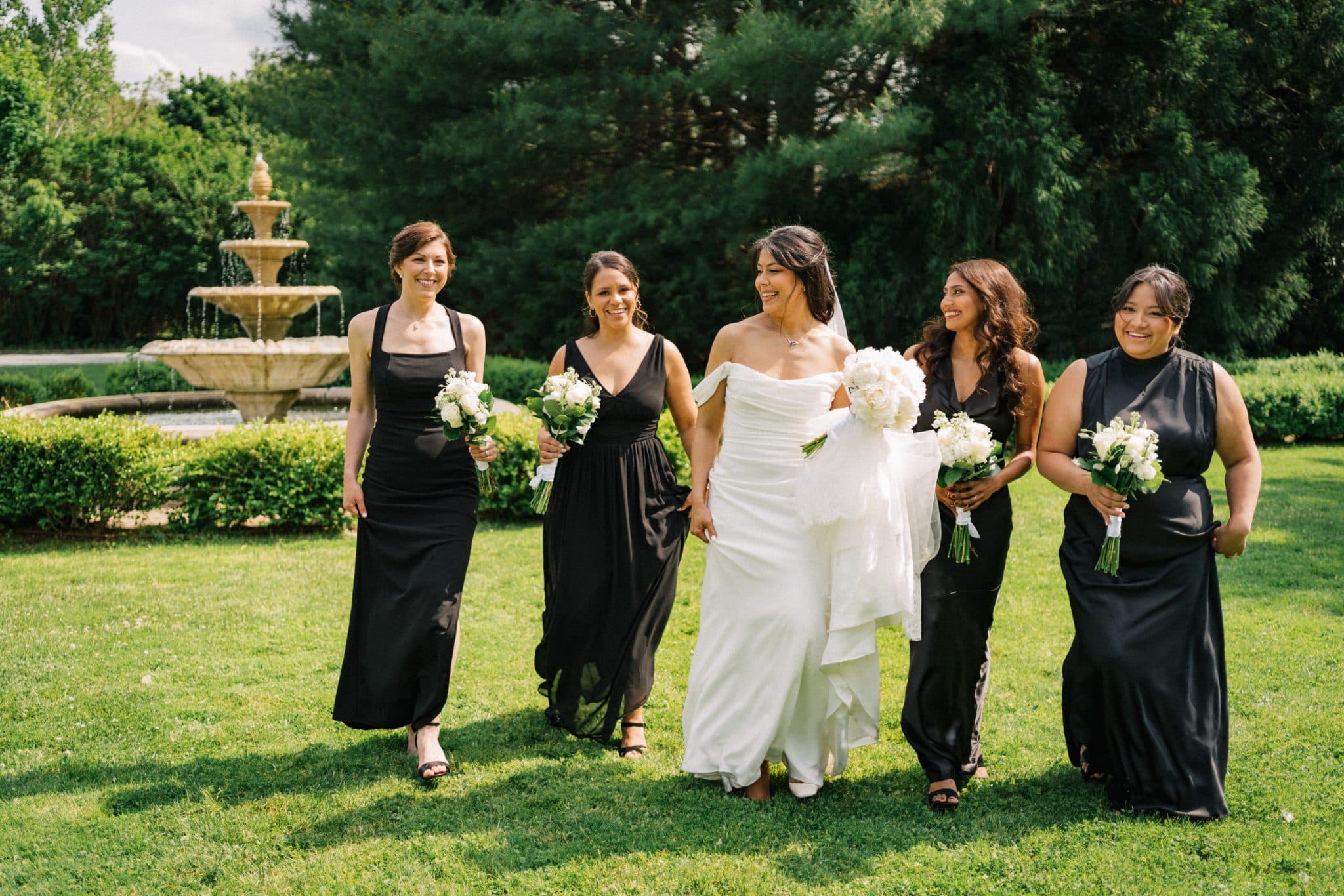A bride in a white dress walks with four bridesmaids in black dresses, holding white bouquets, in front of a fountain and greenery.