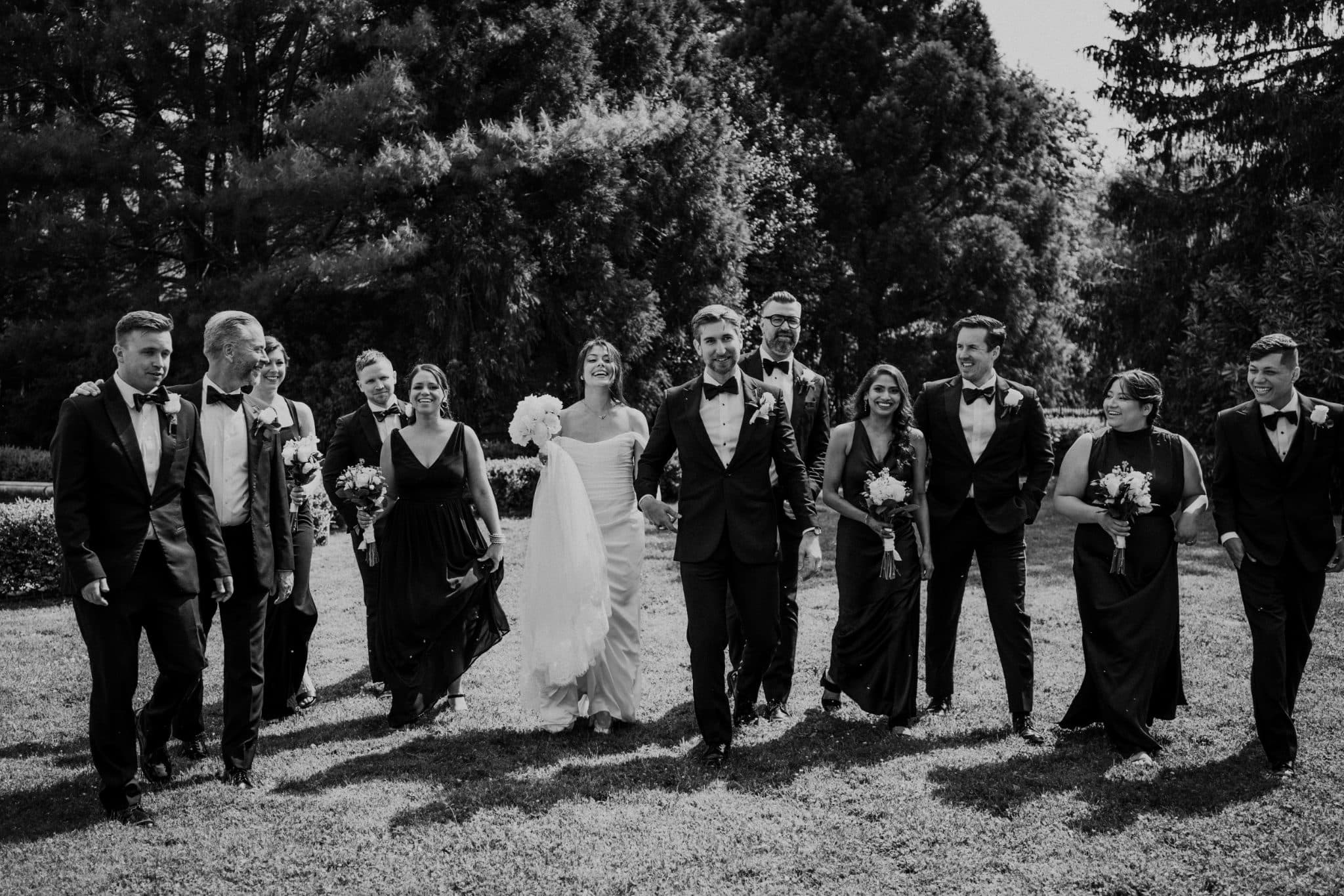 A black and white wedding photograph shows a bride and groom walking with their bridal party in a grassy area surrounded by trees.