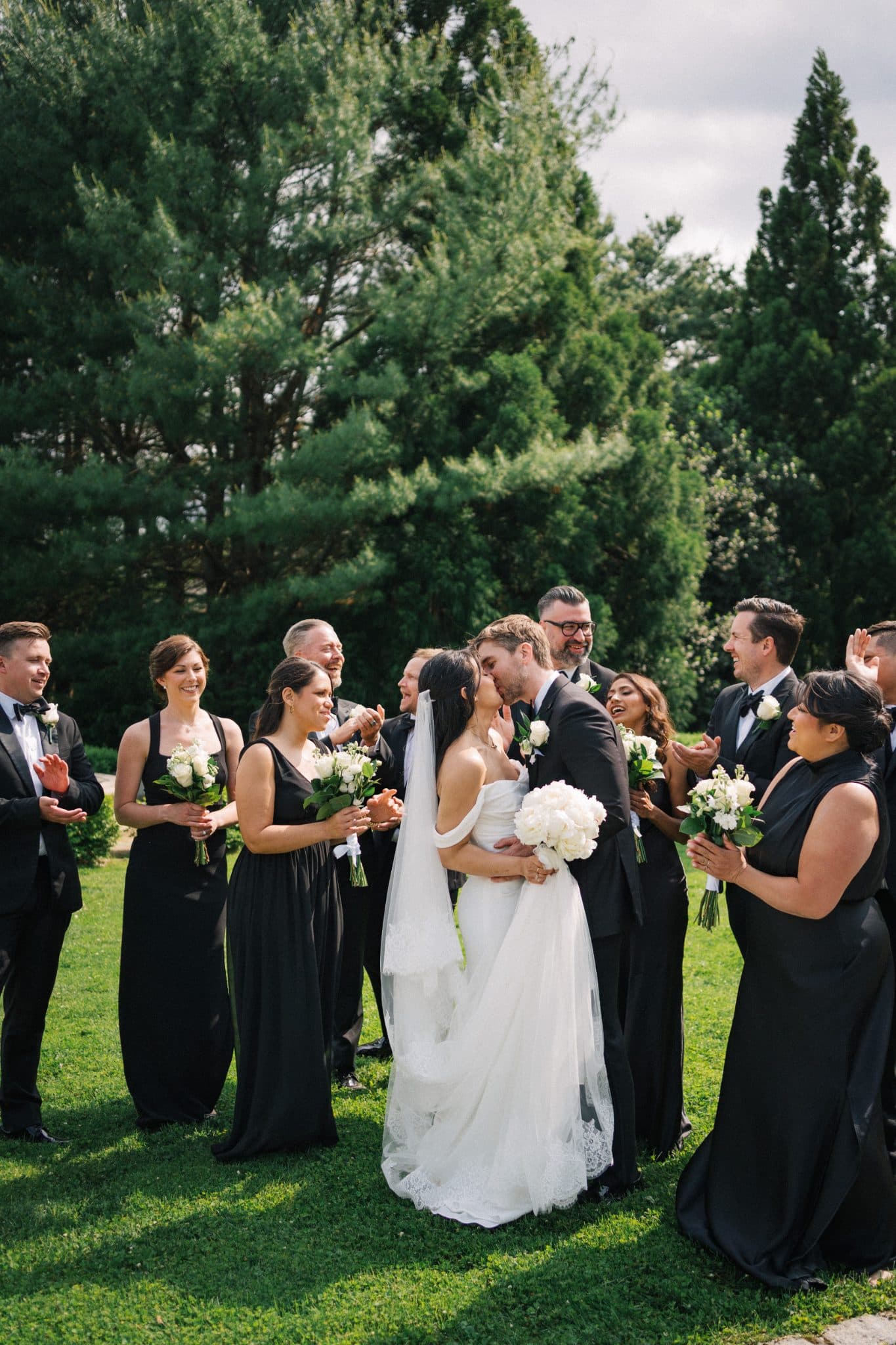 A bride in a white gown kisses a groom in a black suit surrounded by a group of people in black attire holding bouquets.
