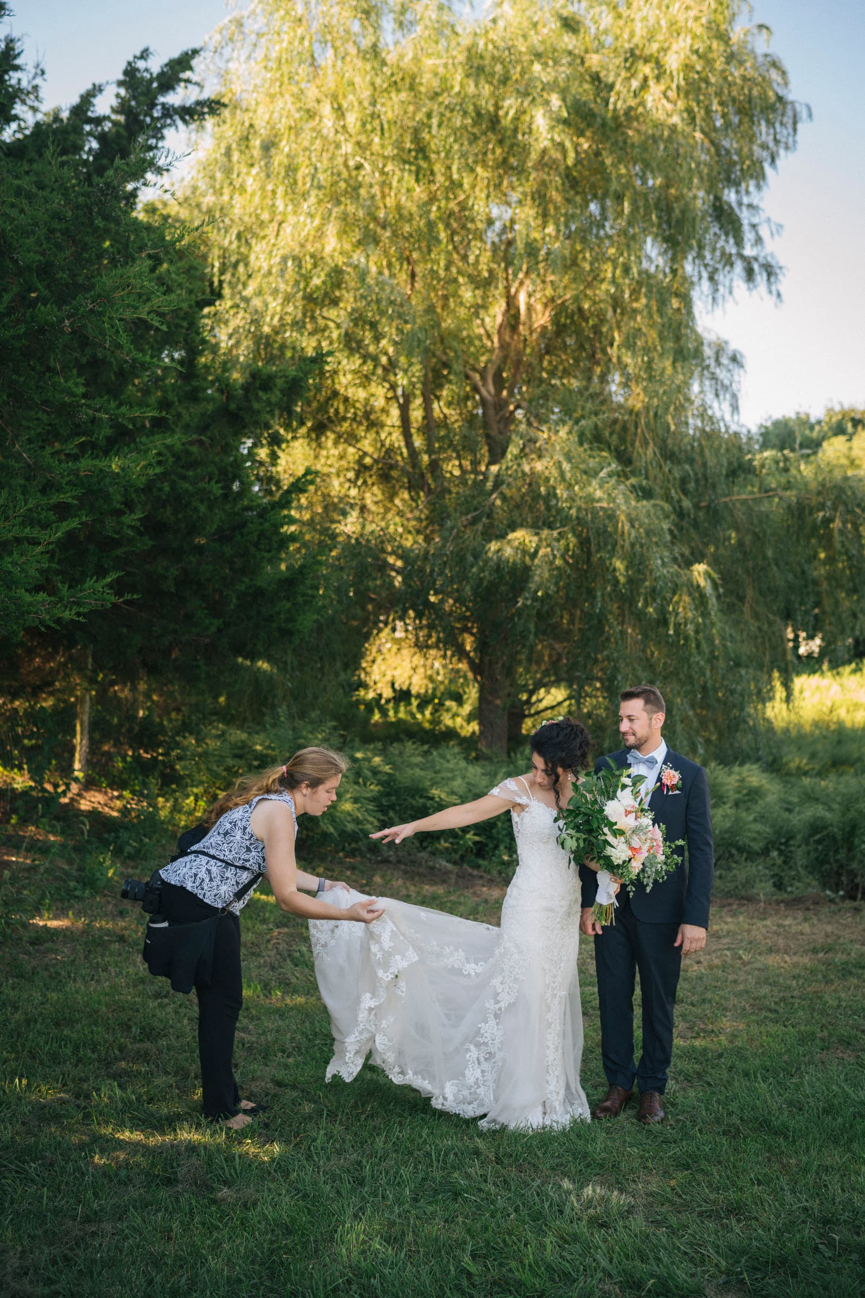 Beth arranging the train of a bride's lace gown while the bride and groom stand together beneath a willow tree