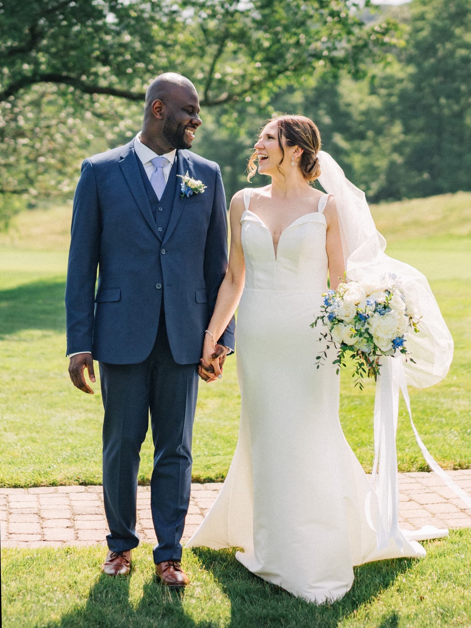 Bride and groom holding hands on a sunlit lawn, smiling at each other as her veil catches the wind