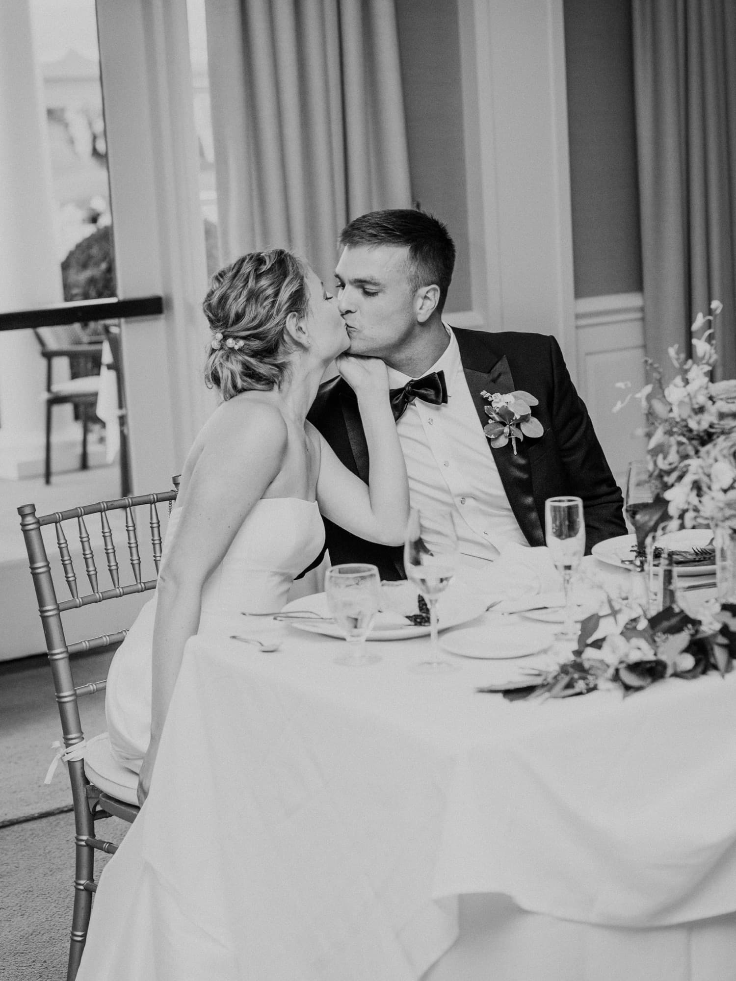 Photograph of a bride and groom kissing at their sweetheart table, champagne flutes and a garden-rose centerpiece in front of them