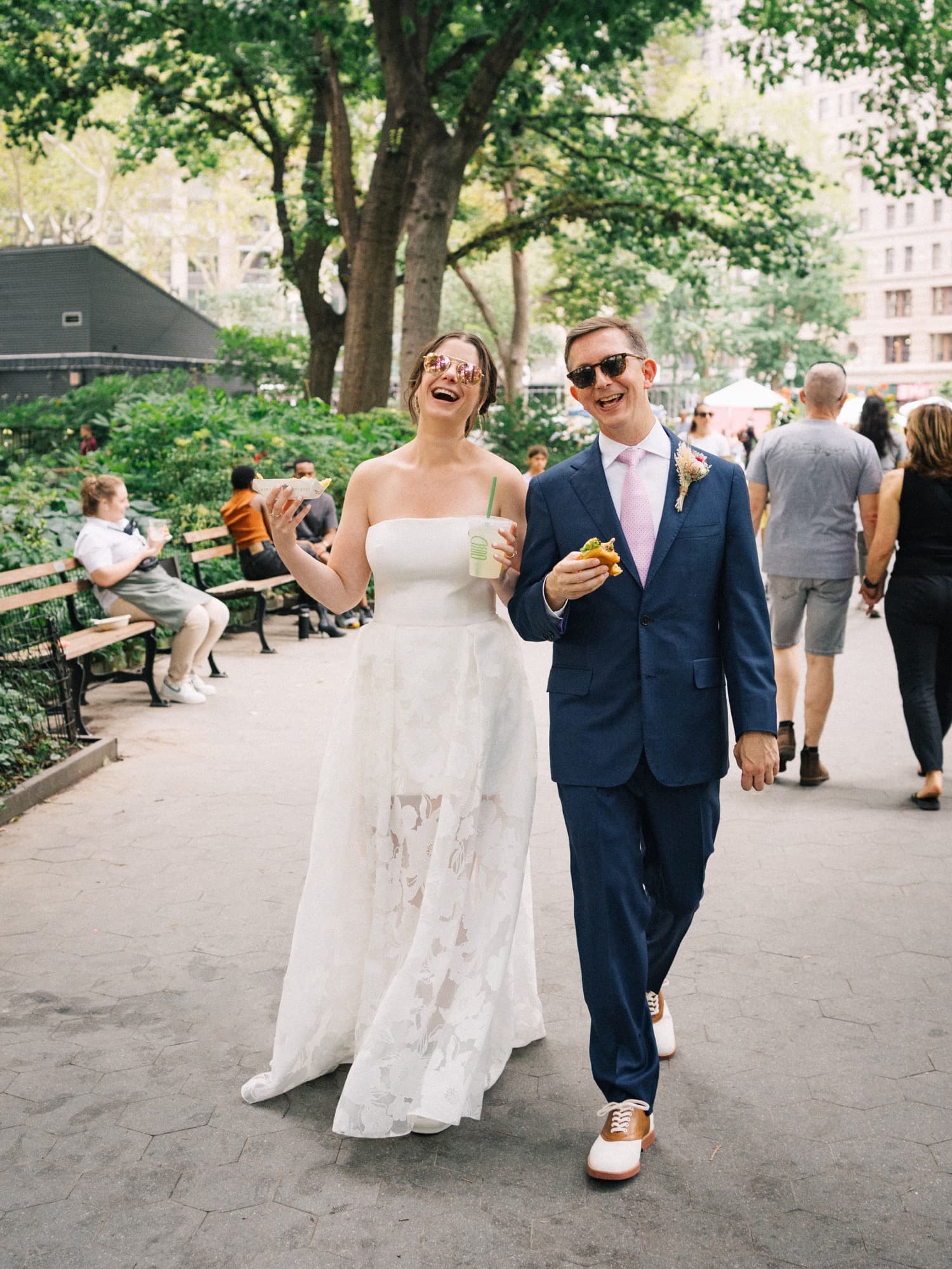 Bride and groom laughing as they walk through a city park in sunglasses, holding fries, a burger, and a lemonade
