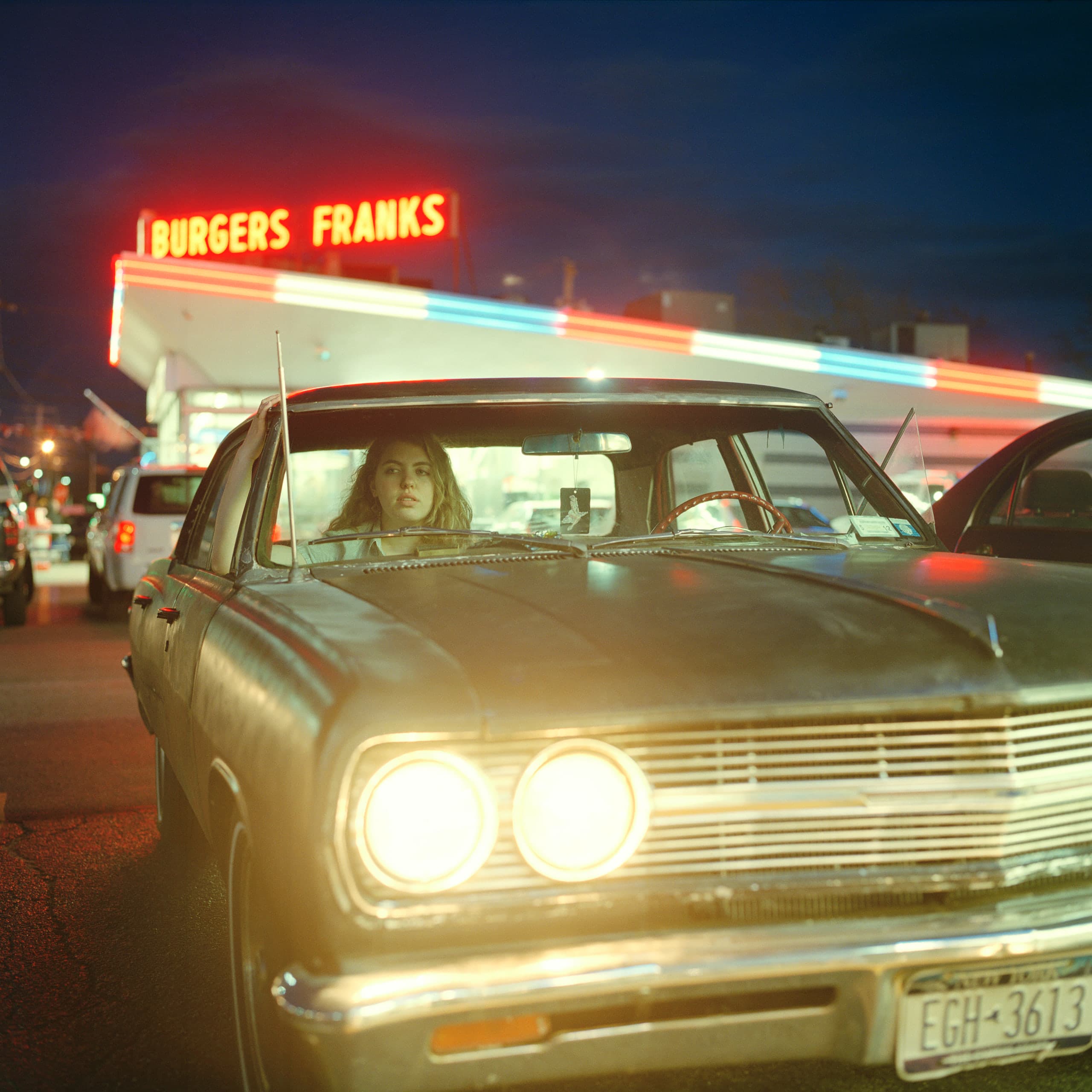 Self-portrait of Beth at dusk behind the wheel of a vintage car, a neon burgers-and-franks sign glowing behind her.