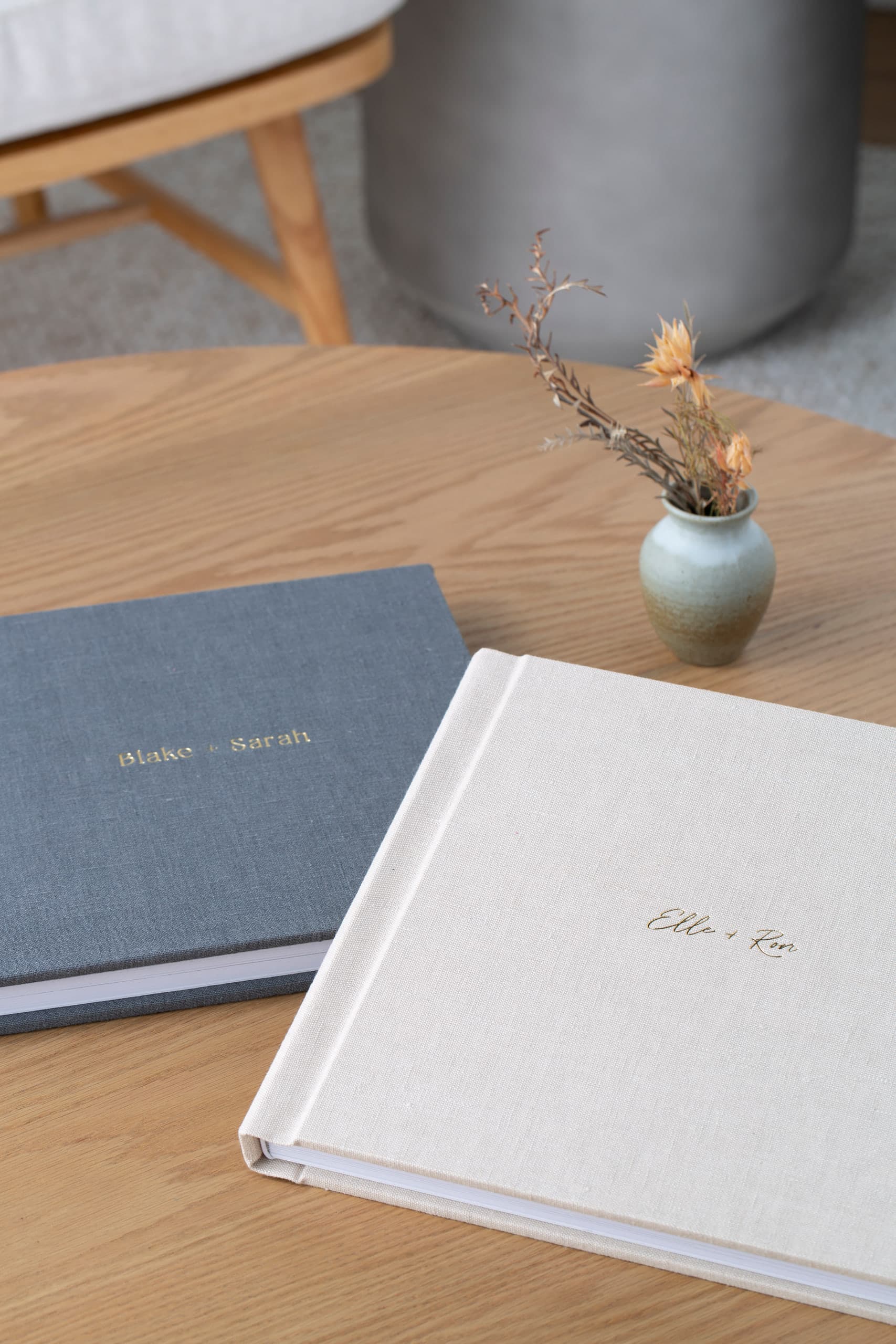 Two linen wedding albums on a wood coffee table — one in slate gray with names debossed in gold, one in ivory with a script title — beside a small vase of dried flowers