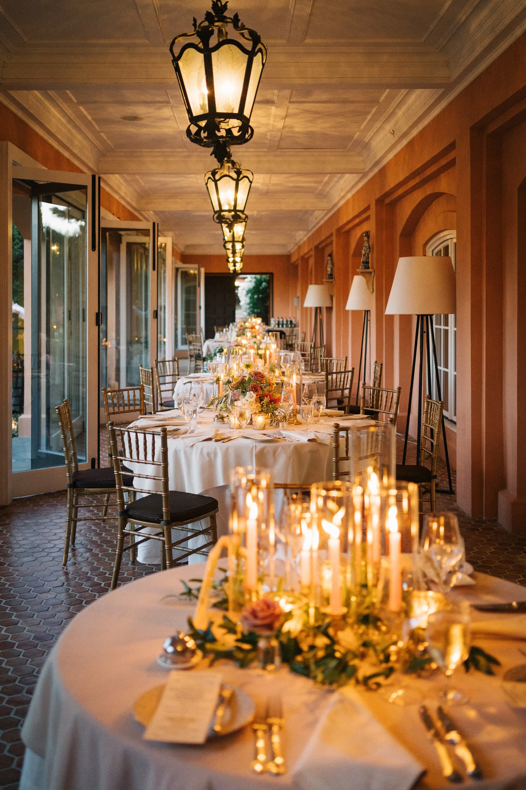 Candlelit round tables running the length of a terracotta loggia under iron lanterns
