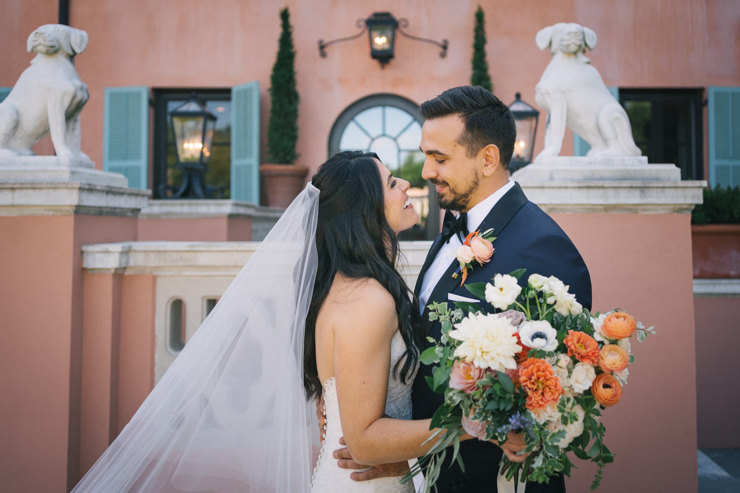 Bride and groom smiling at each other outside a pink villa