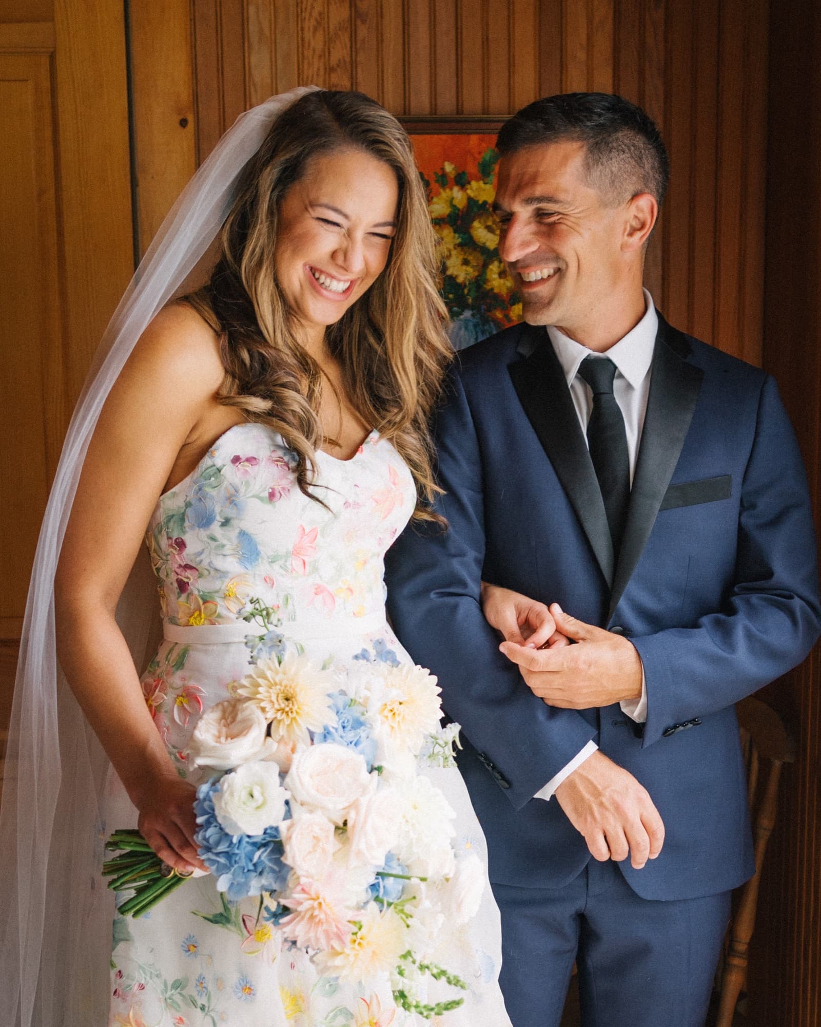 Sarah laughing beside Kevin in a wood-panelled room, holding a pastel bouquet in a hand-painted floral gown