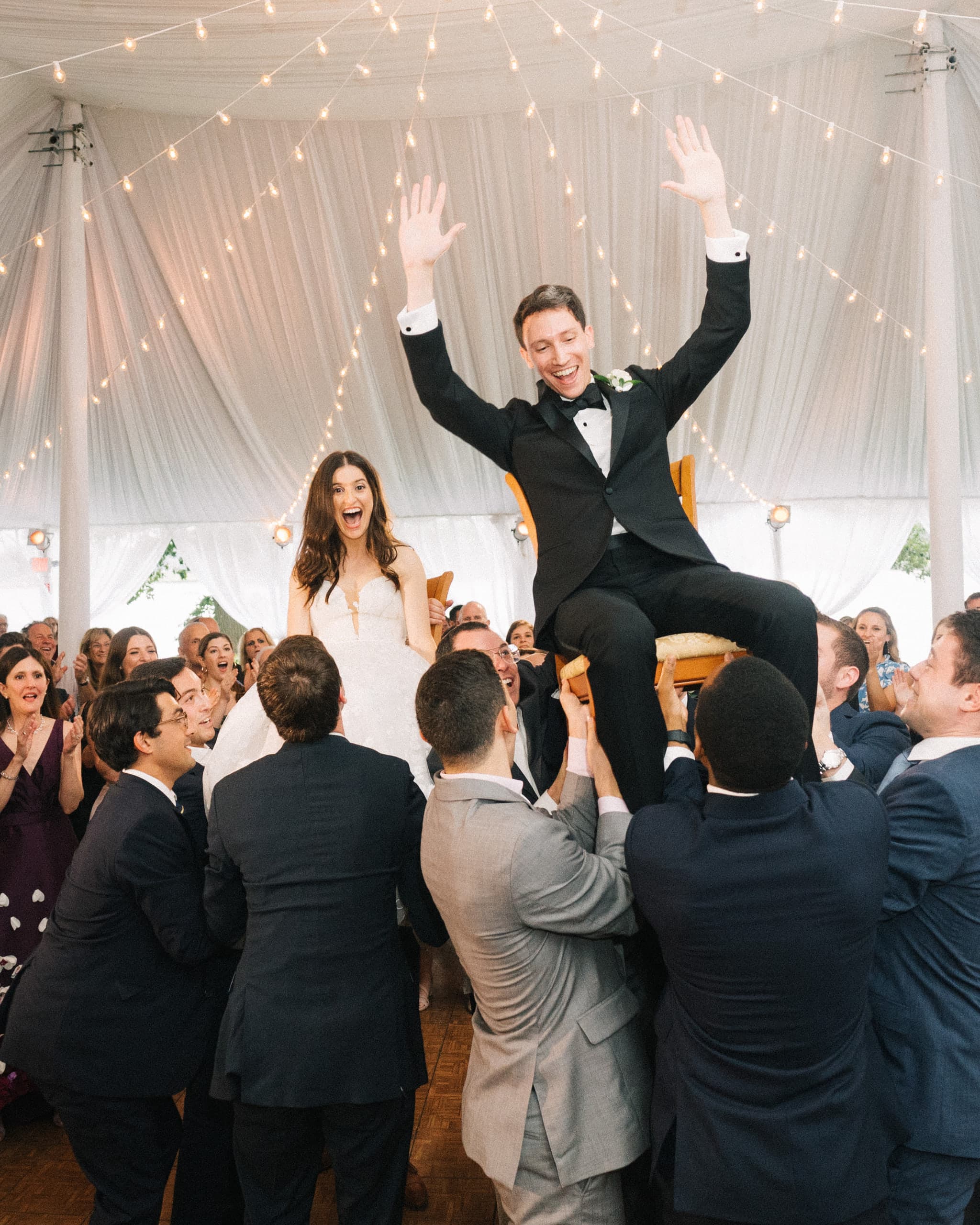 Bride and groom lifted on chairs during the hora beneath a string-lit tent
