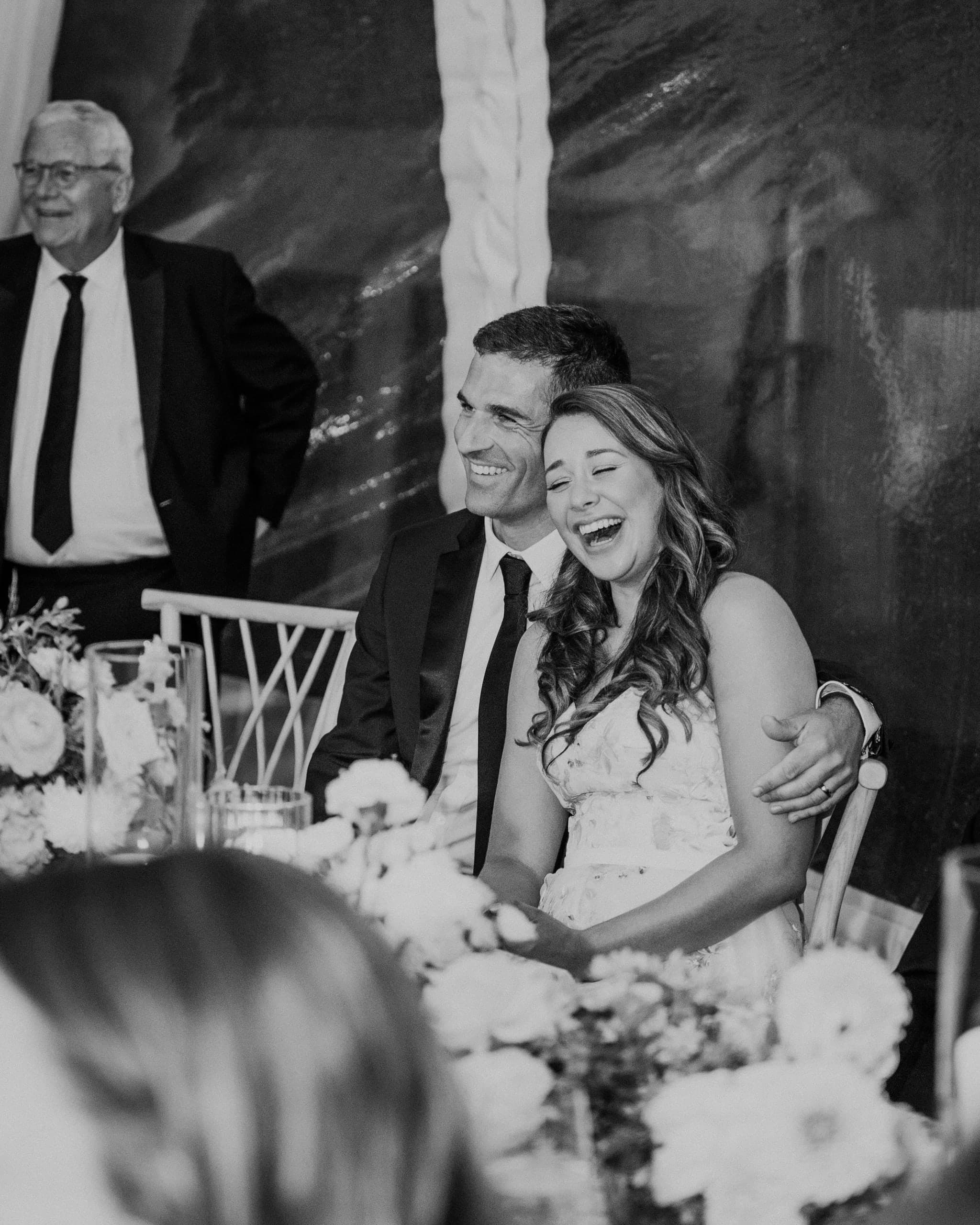 Bride and groom laughing together at their reception table, in black and white