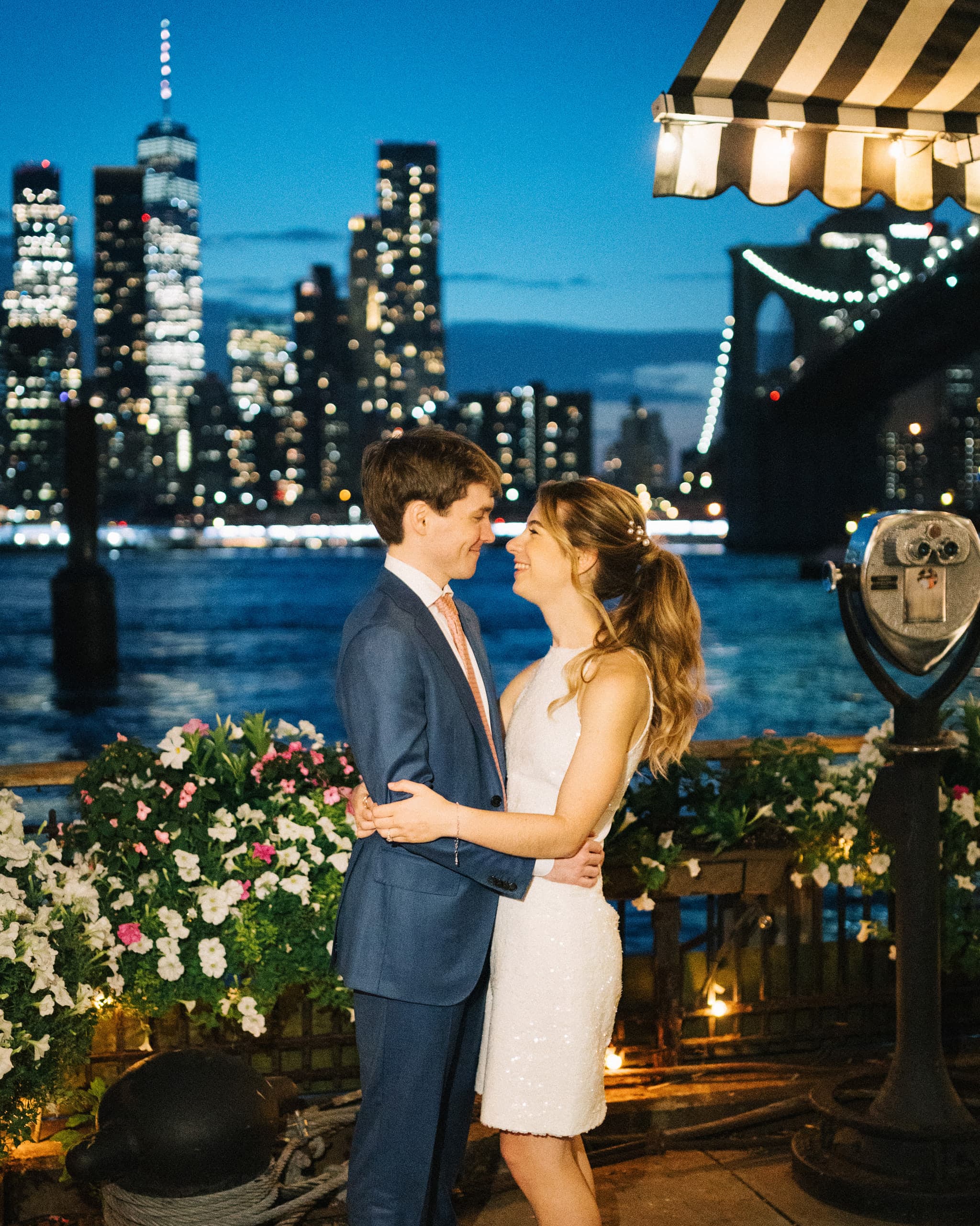 Couple embracing on a waterfront terrace at dusk, the Manhattan skyline and Brooklyn Bridge behind them