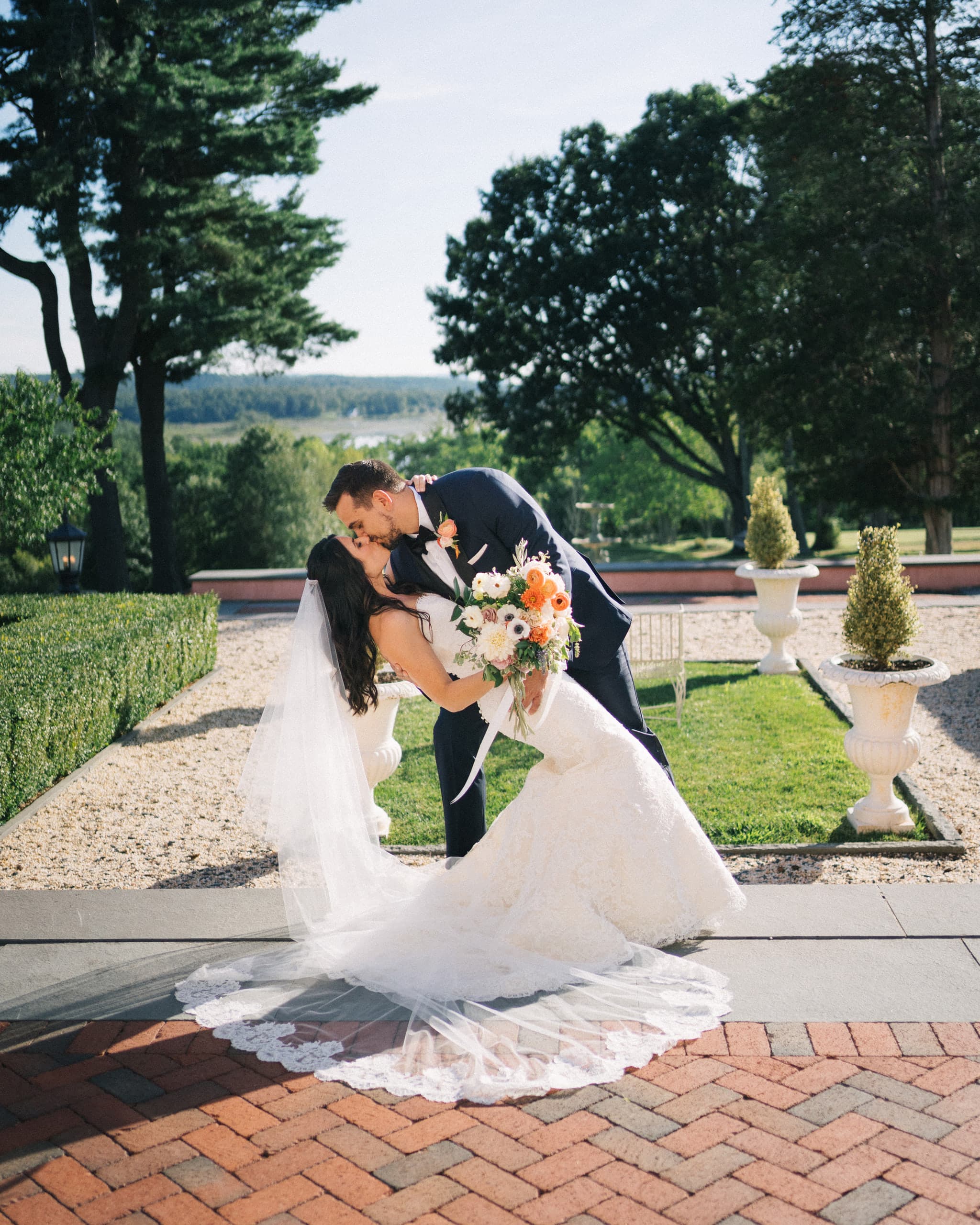 Groom dipping the bride for a kiss on a garden path, her lace train fanned across the brick