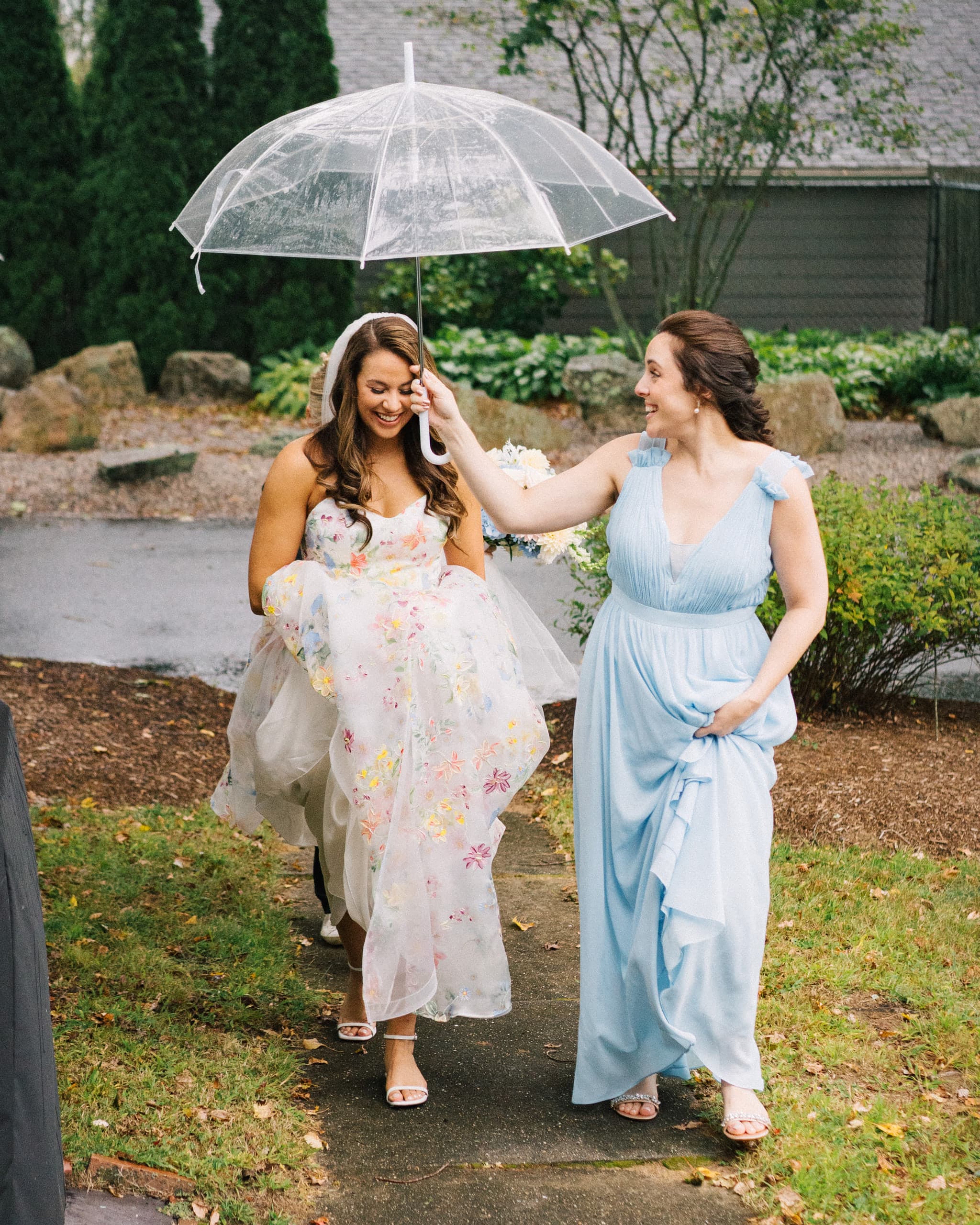Bride in a floral gown walking a garden path while a bridesmaid holds a clear umbrella over her