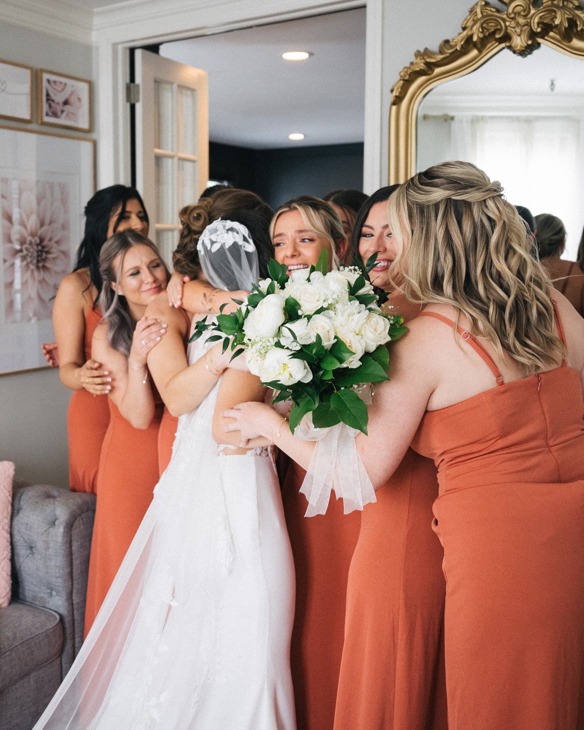 Bridesmaids in rust dresses embracing the bride, who holds a white rose bouquet