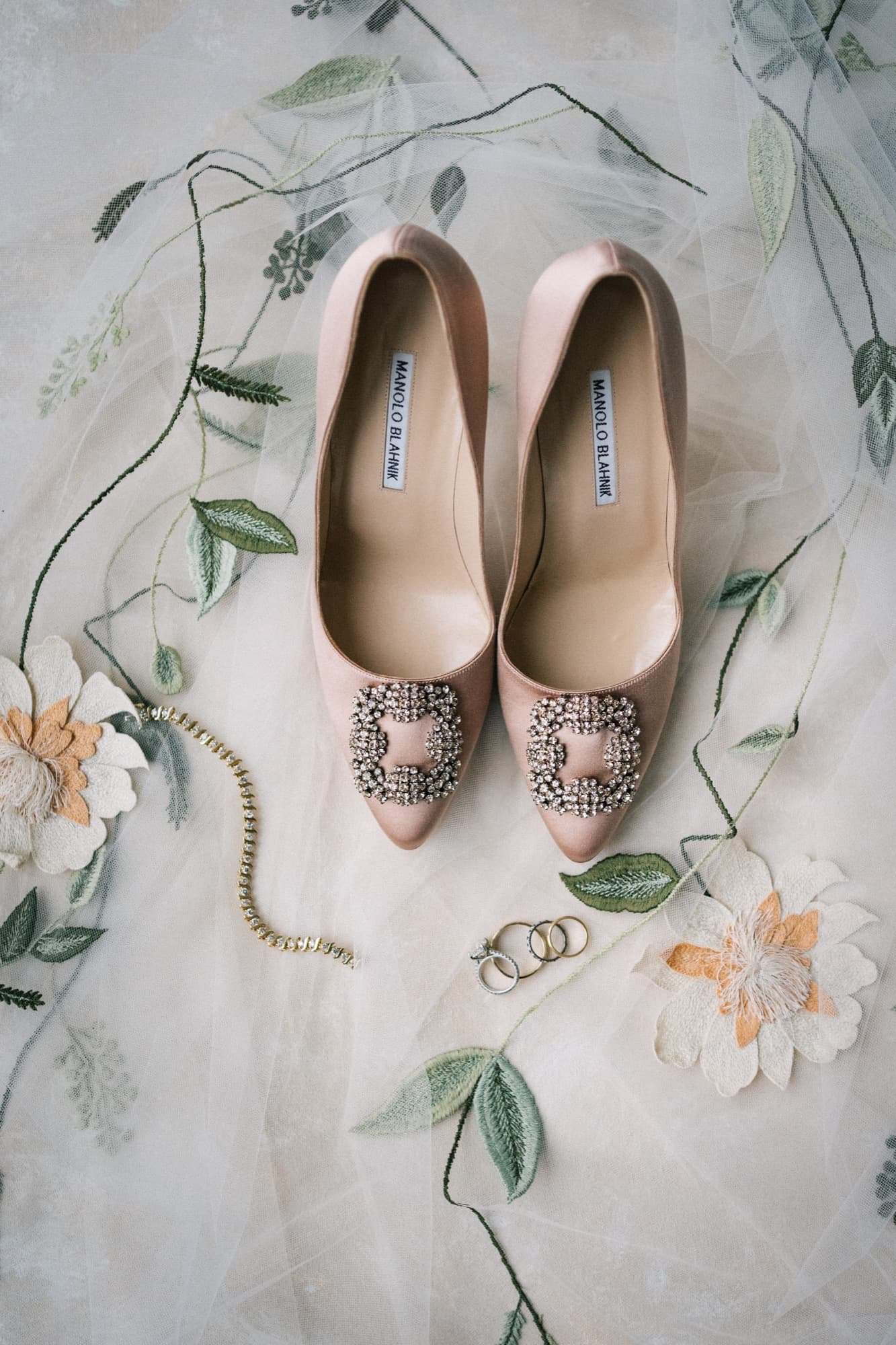 Blush satin bridal heels with jeweled buckles beside rings and a bracelet on an embroidered veil
