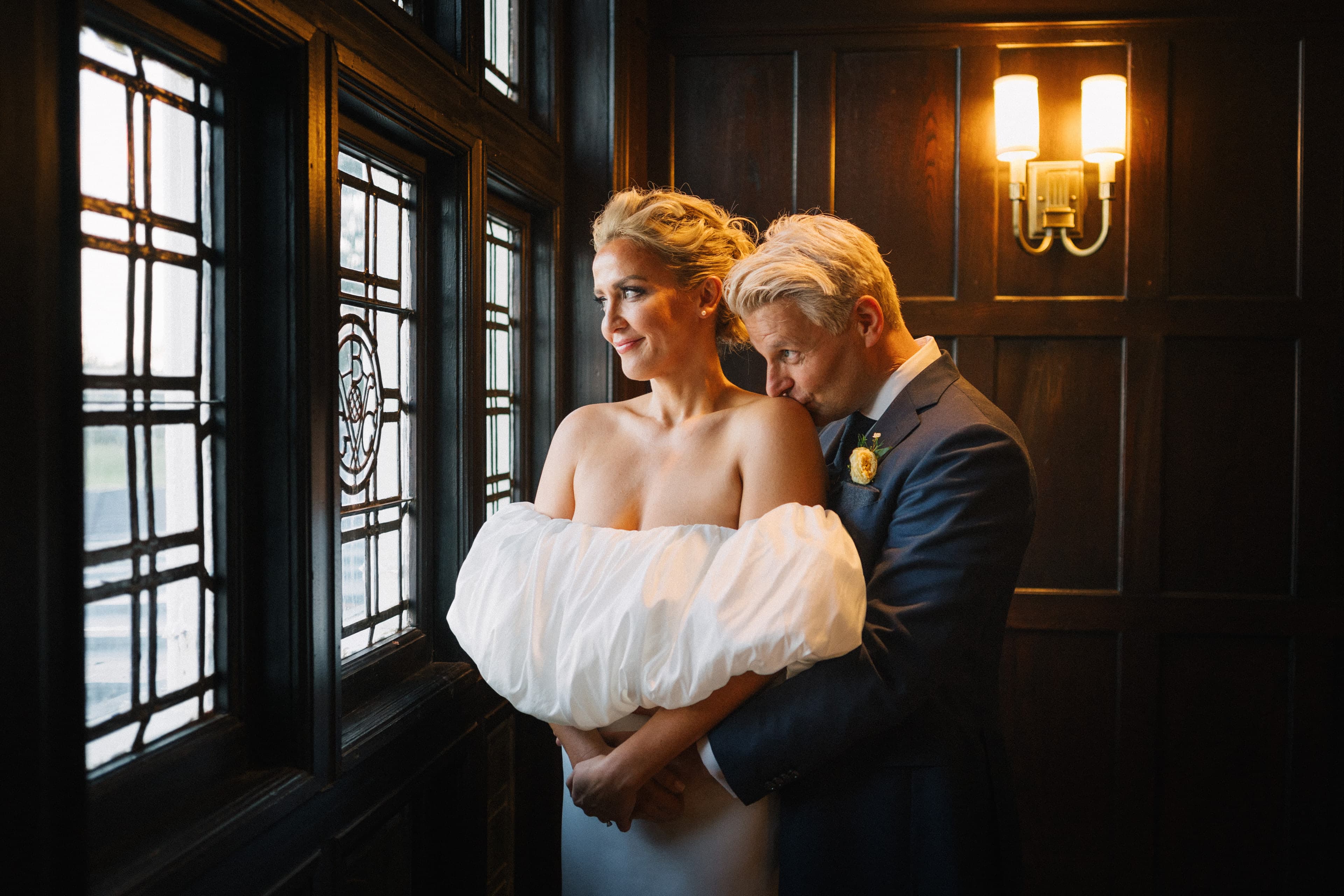 An embrace by the leaded-glass windows in a wood-paneled room, lit by a wall sconce