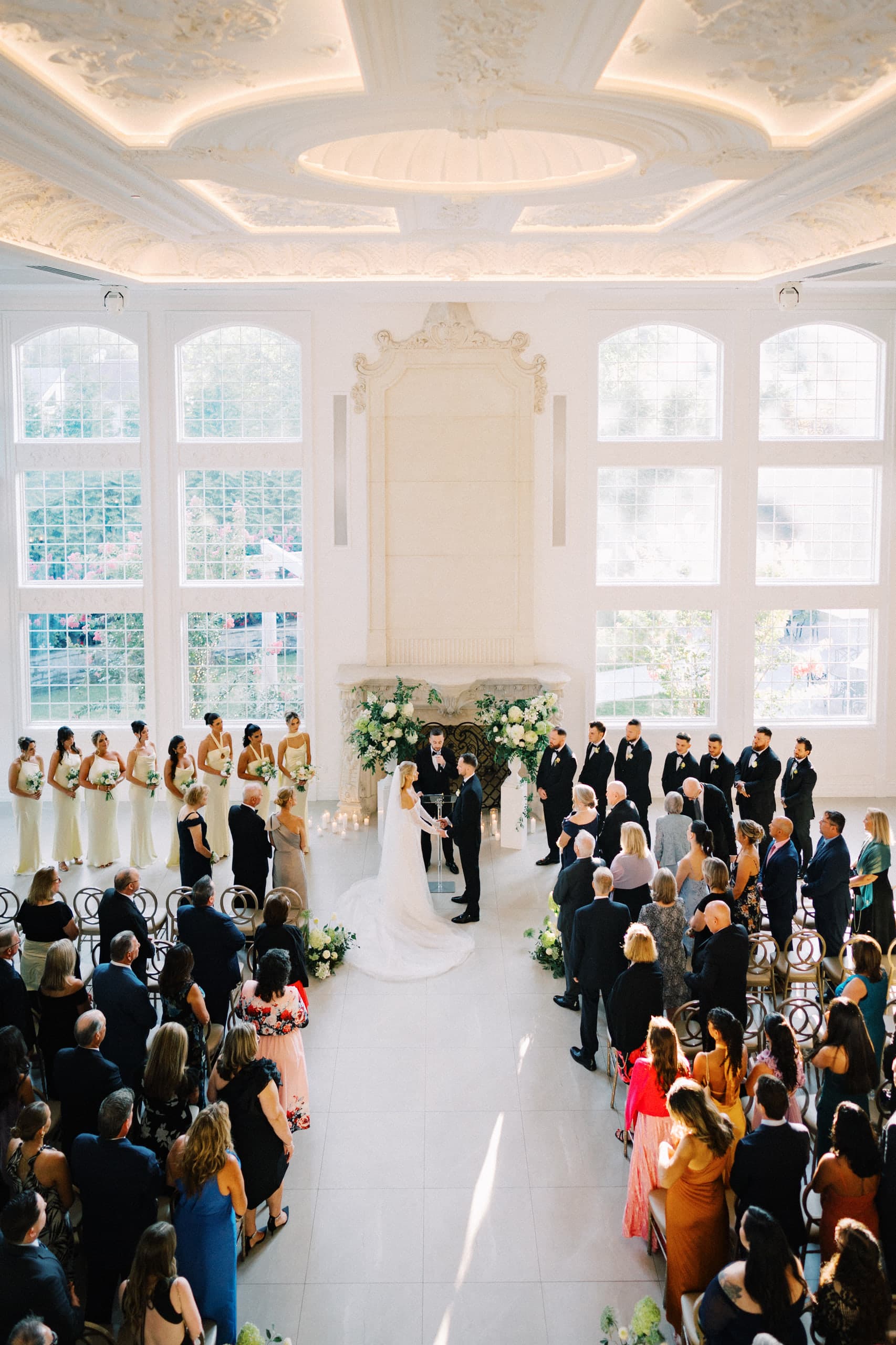 Vows before the fireplace of a sunlit white hall, seen from above