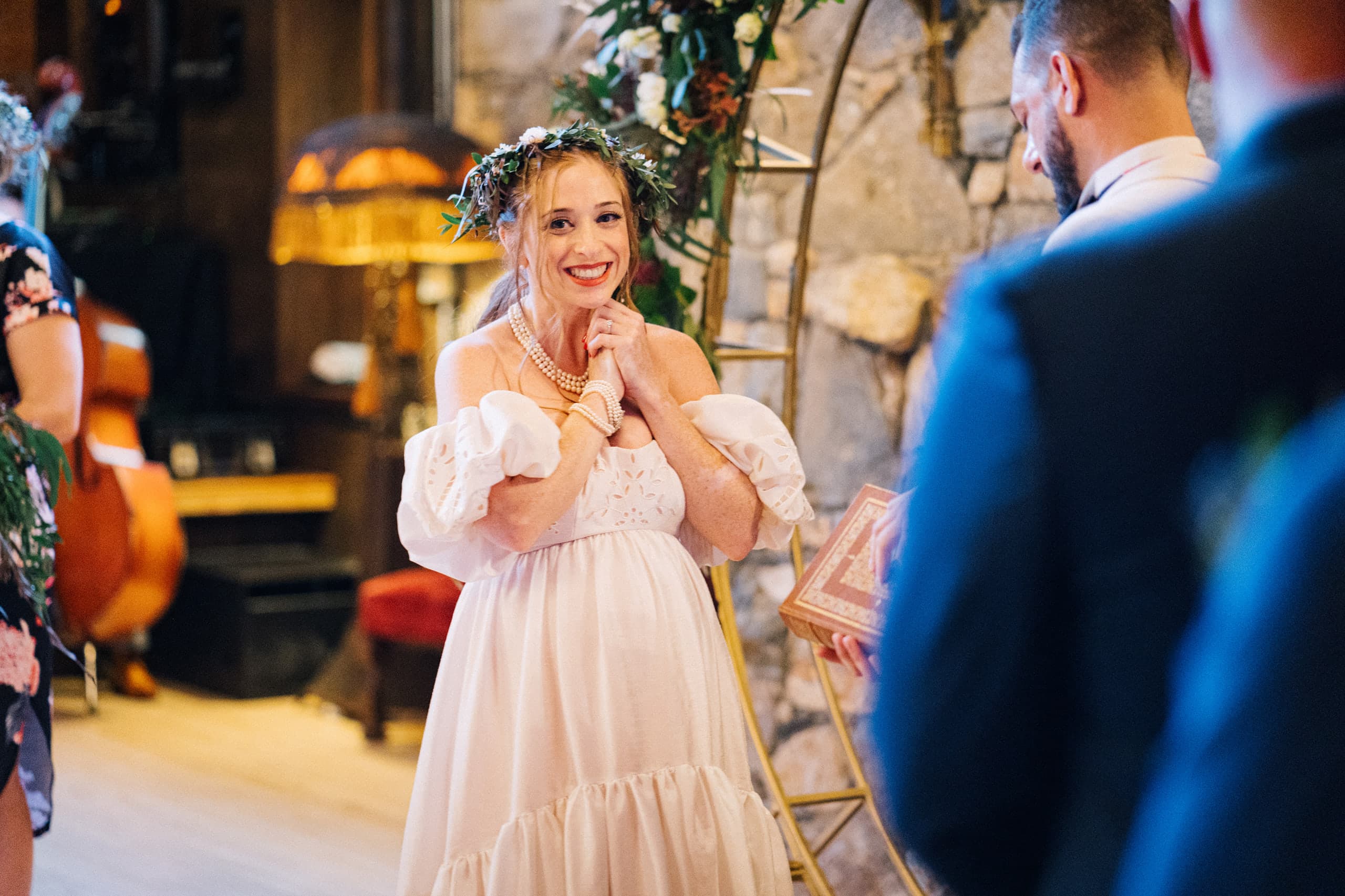 A flower-crowned bride clasps her hands, beaming at her groom by the stone wall