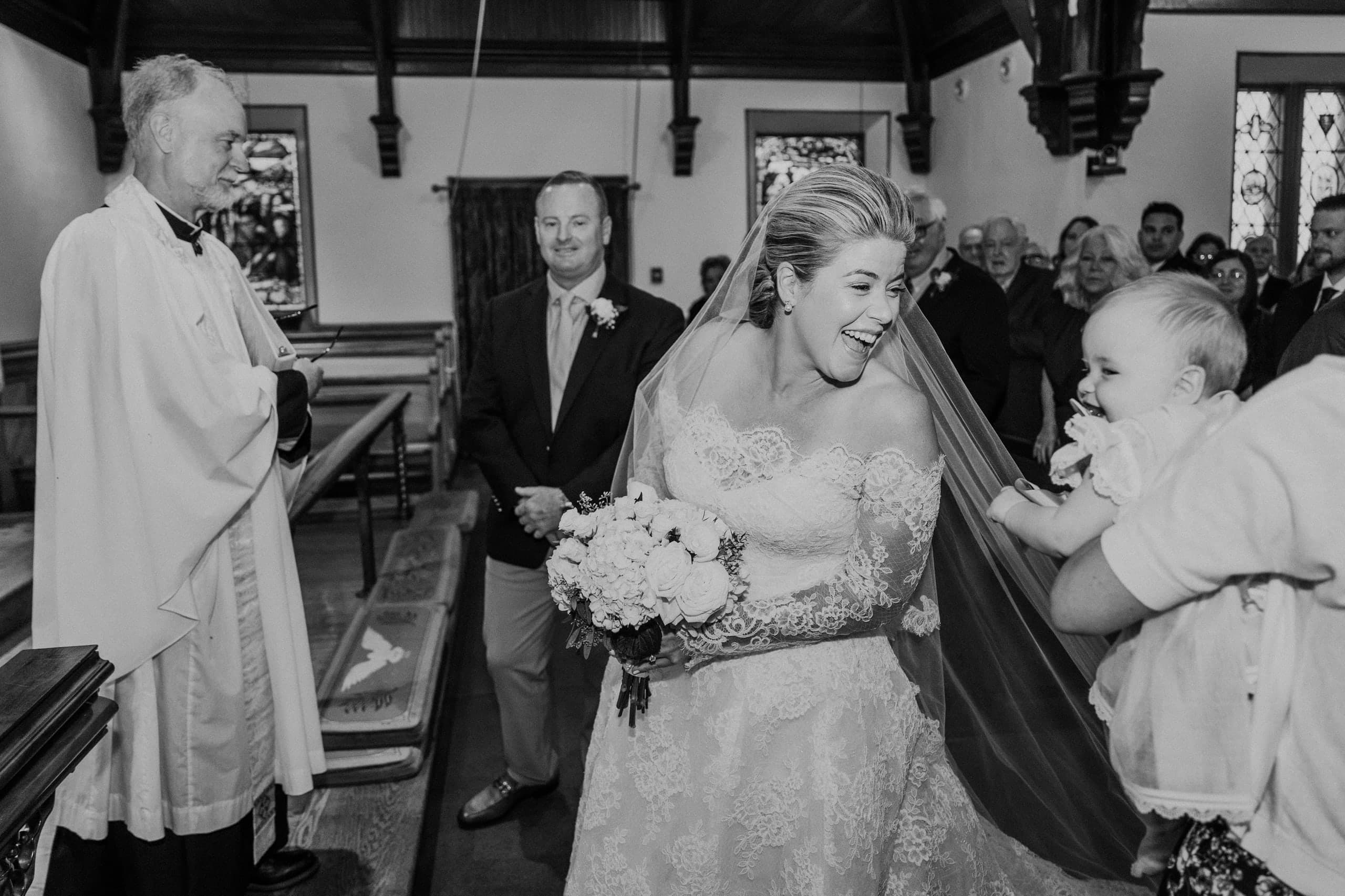 The bride laughing down the aisle as a baby reaches for her veil, black and white