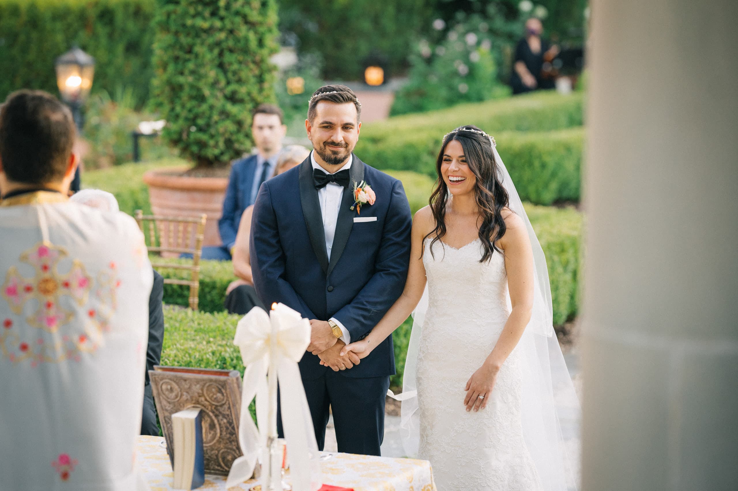 Smiles across joined hands at a garden ceremony, priest's robes in the foreground
