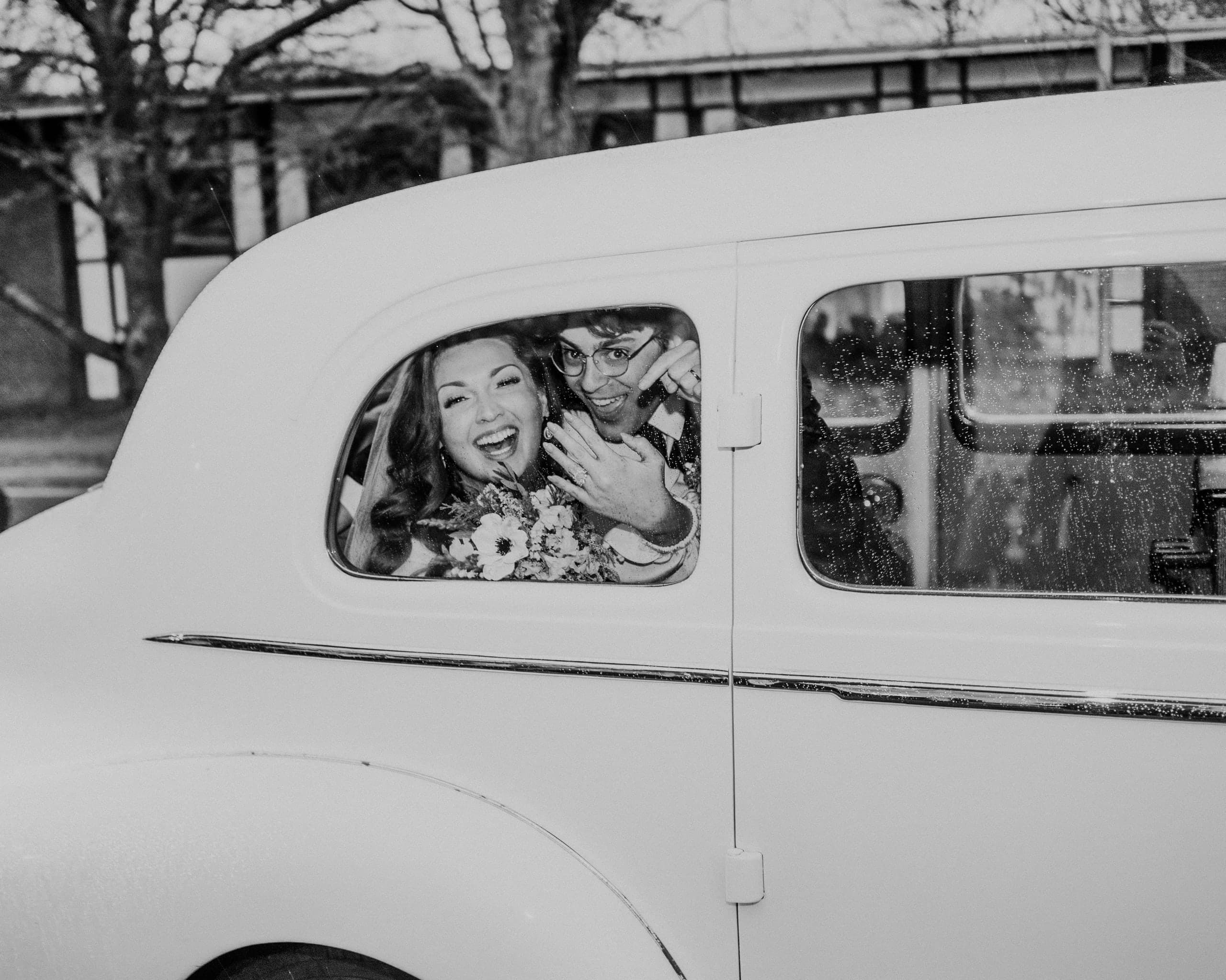 The newlyweds mugging from the window of a vintage white car, black and white